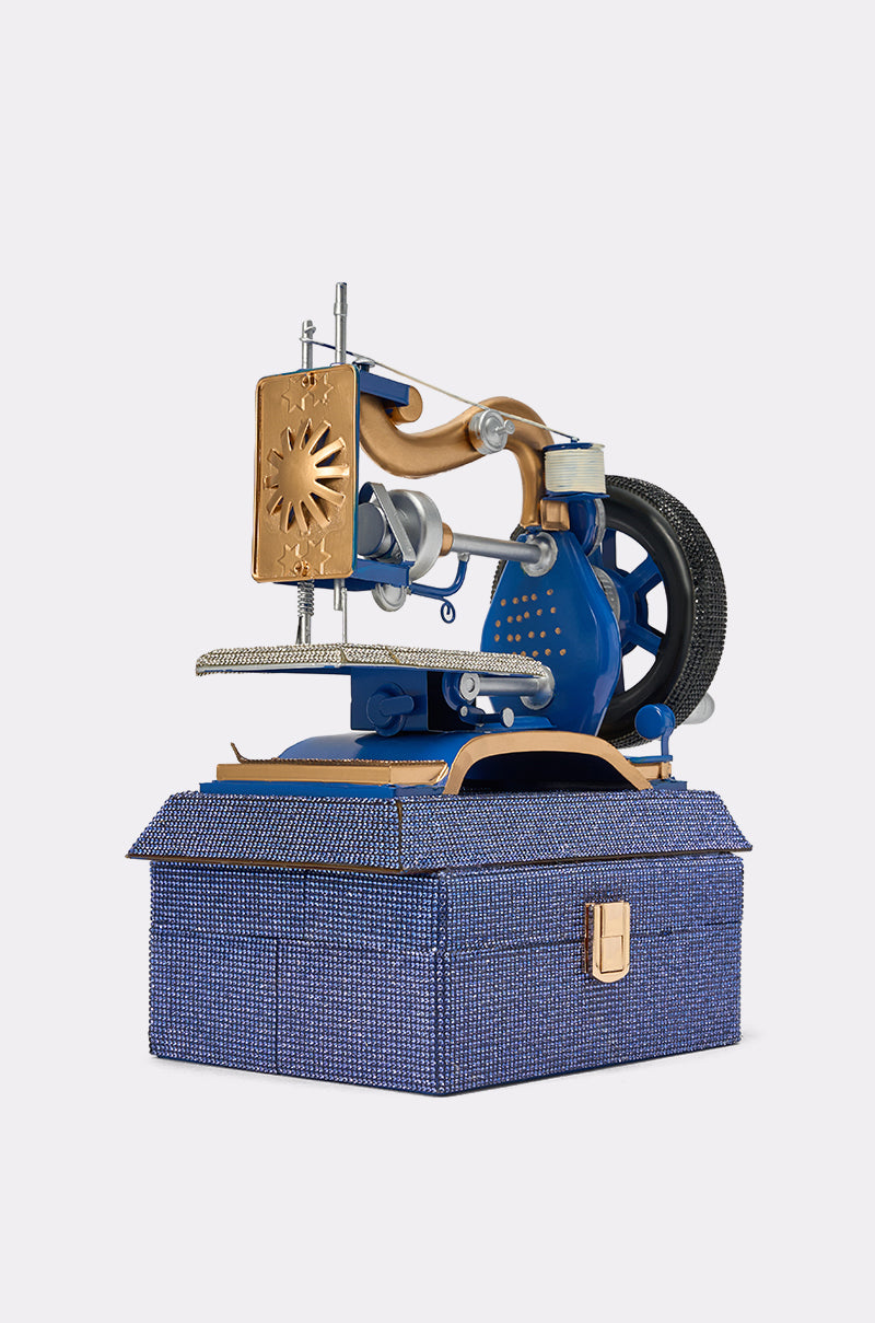 The SEW IT BAG, a vintage-inspired blue and gold sewing machine bag, rests on a textured blue box with a handle and clasp, resembling a charming novelty handbag against a plain light background. 3