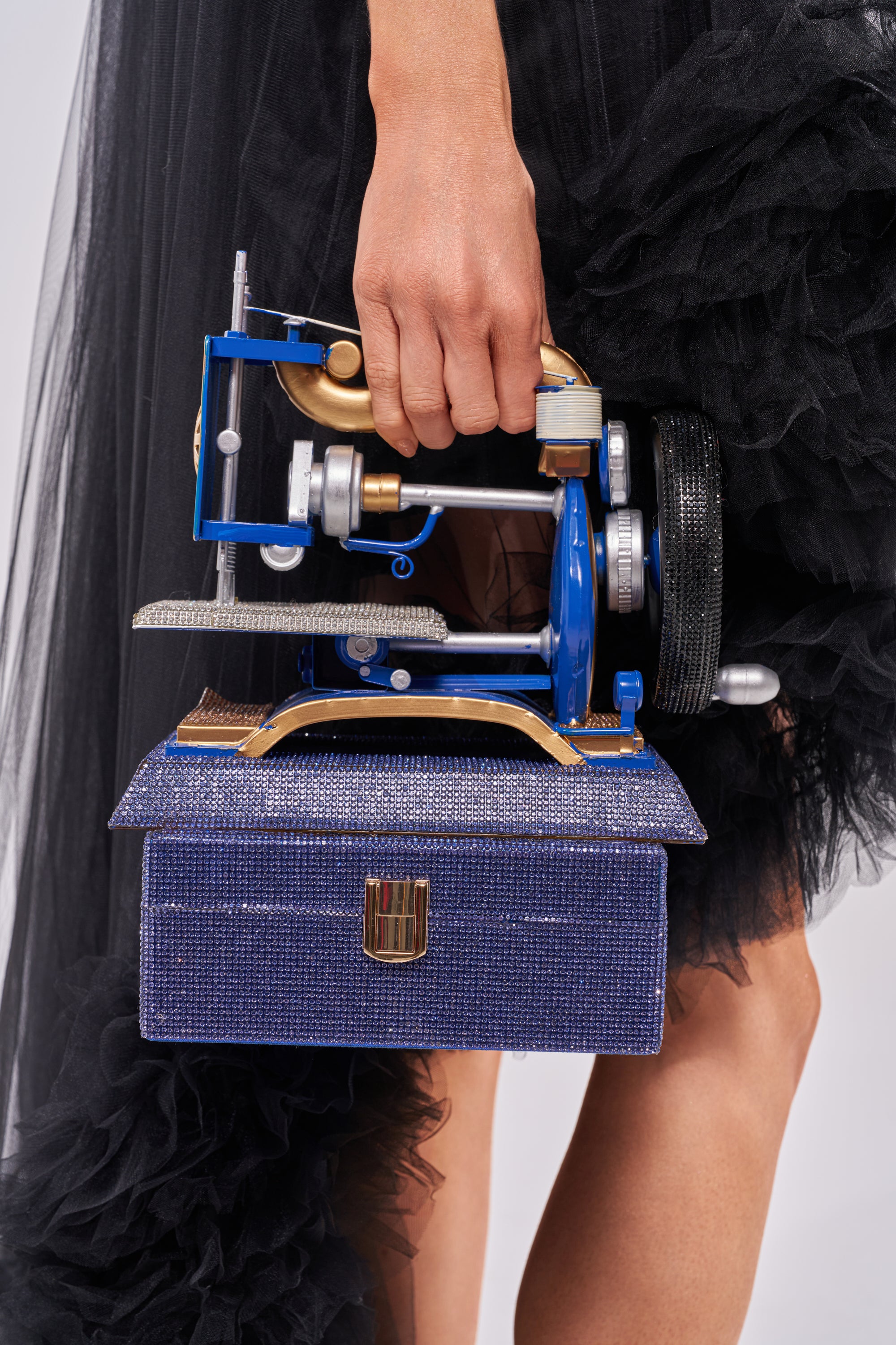 A person in a black tulle dress holds the SEW IT BAG, a novelty handbag shaped like a vintage sewing machine with blue, gold, and silver accents and a textured, boxy base. 1