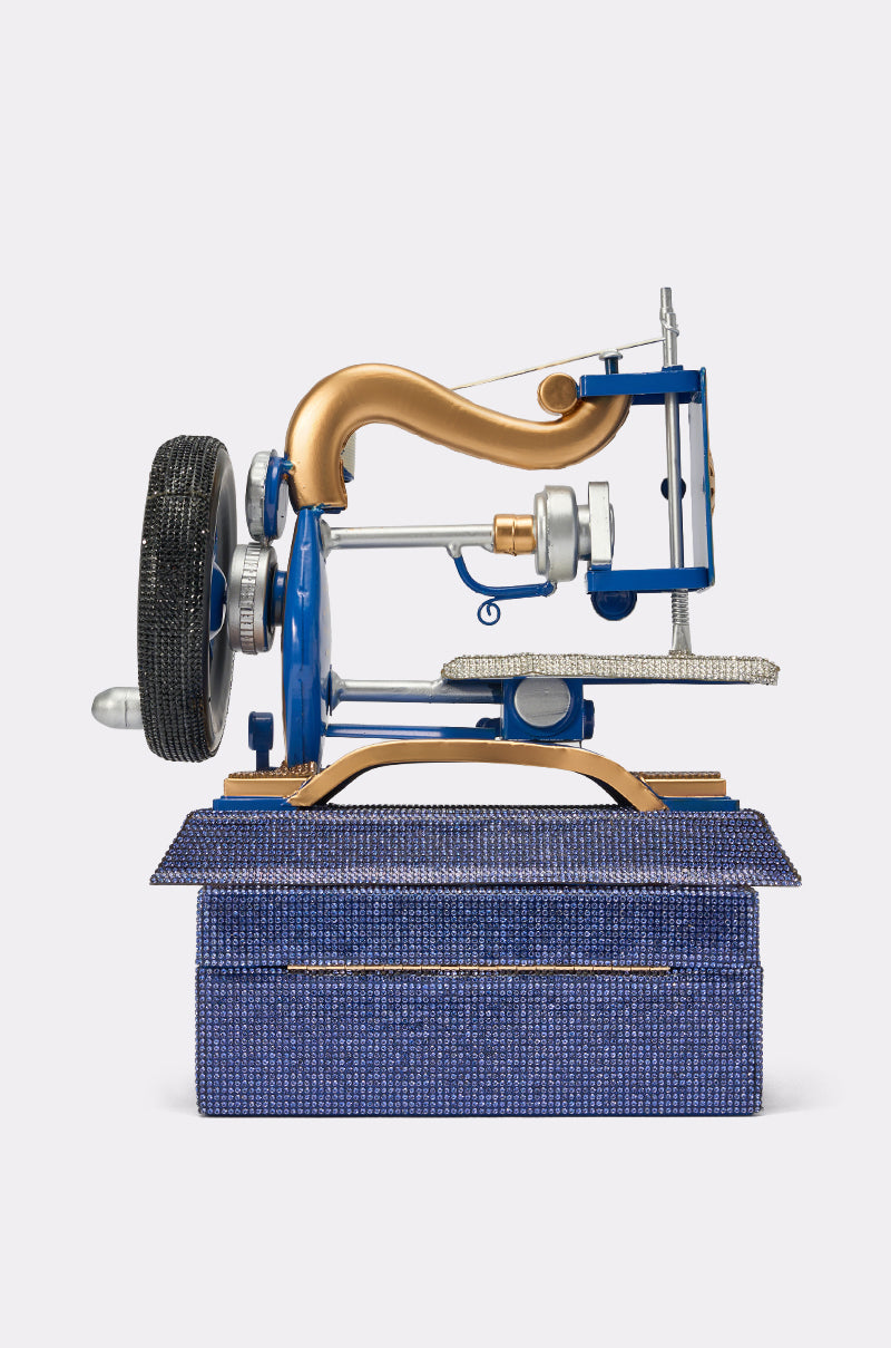 The SEW IT BAG, designed as a blue and gold vintage sewing machine reminiscent of a rhinestone purse, sits on a sparkling blue rectangular box against a plain white background. 4