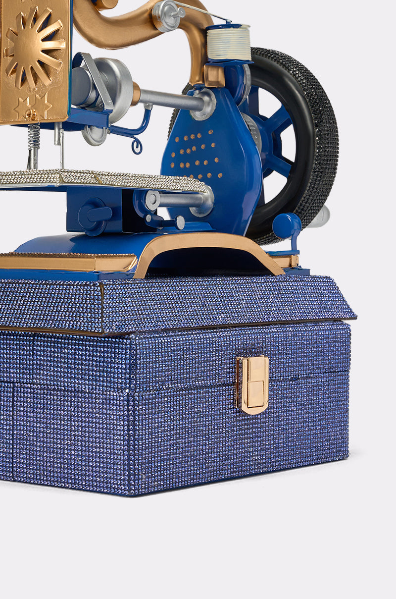 A close-up of the SEW IT BAG, a blue textured vintage sewing machine handbag with a gold clasp and handle, topped with intricate gold, silver, and blue mechanical components such as wheels and gears. 5