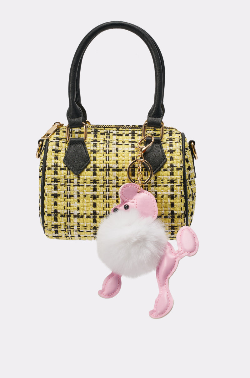 The MIA TWEED MINI PURSE is a small yellow tweed barrel bag with black handles, gold hardware, a crossbody strap, a white fluffy pom-pom, and a pink poodle charm on the handle. 1