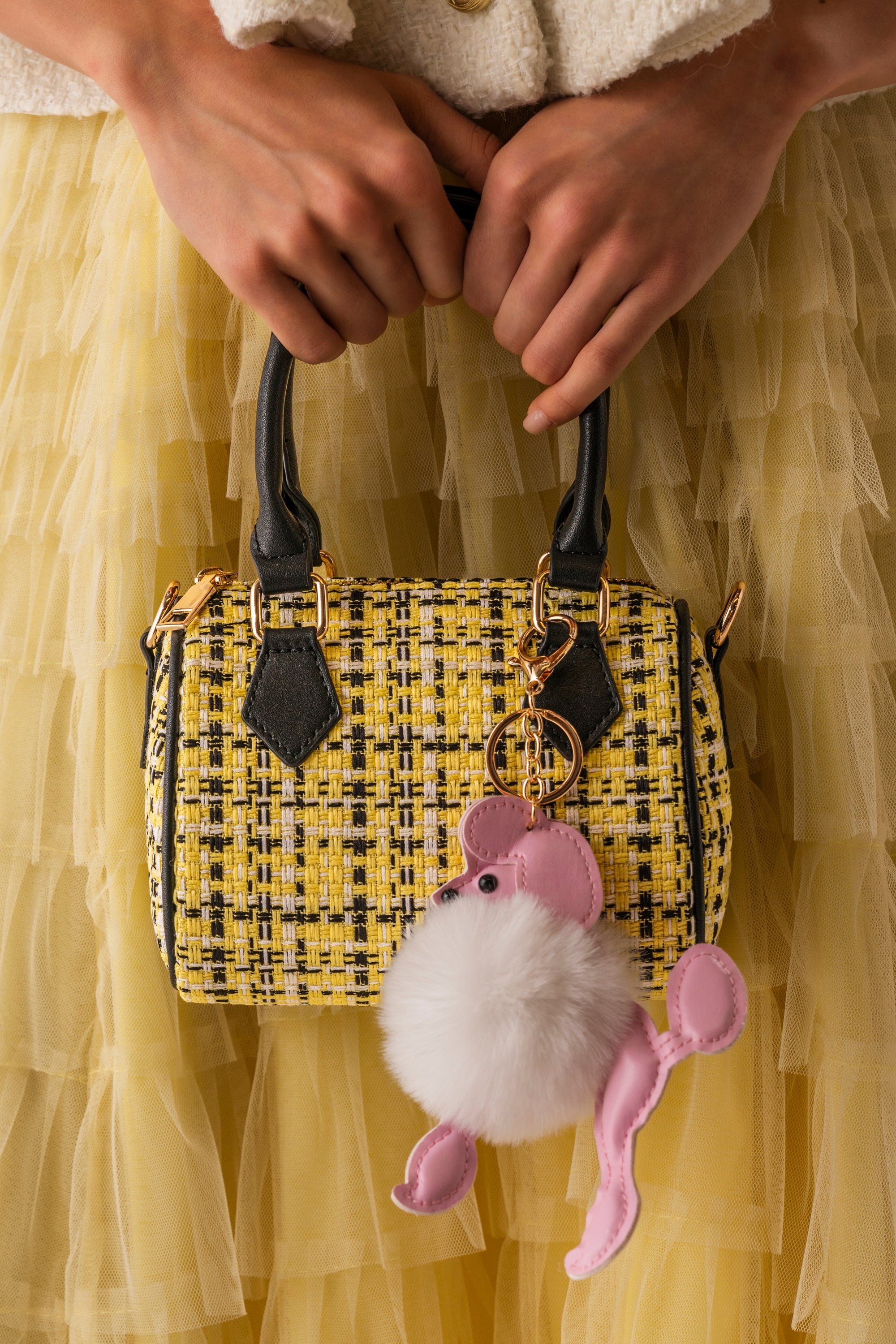 A person holds the MIA TWEED MINI PURSE, a yellow tweed mini barrel bag with black handles and a crossbody strap, accessorized with a fluffy pink and white poodle charm. They pair it with a yellow tulle skirt and light textured top. 0