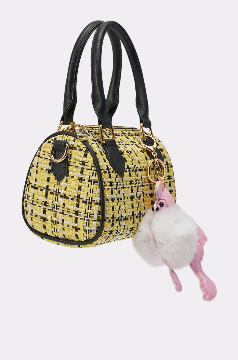 The MIA TWEED MINI PURSE is a small yellow and black barrel bag with black handles and gold hardware, featuring a fluffy white and pink flamingo keychain and a playful poodle bag charm clipped to one handle. 2