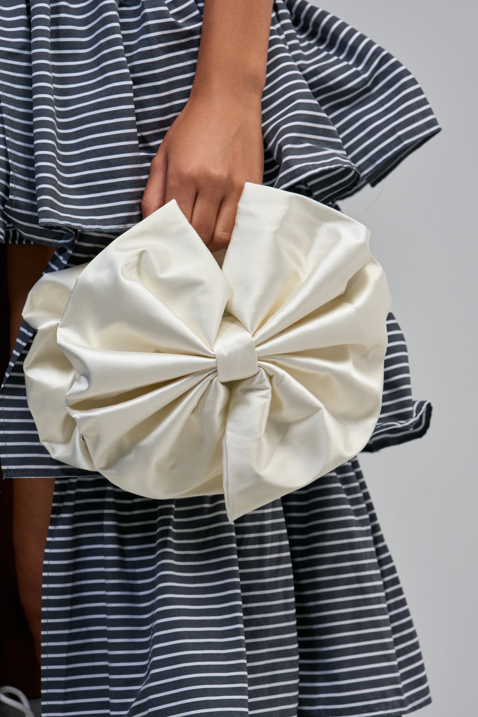 Clutching the WITH A BOW PURSE—an elegant, ivory satin bag beautifully shaped like a delicate bow—a person in a gray and white striped dress showcases the perfect accessory for brides or anyone seeking a sophisticated evening bag.