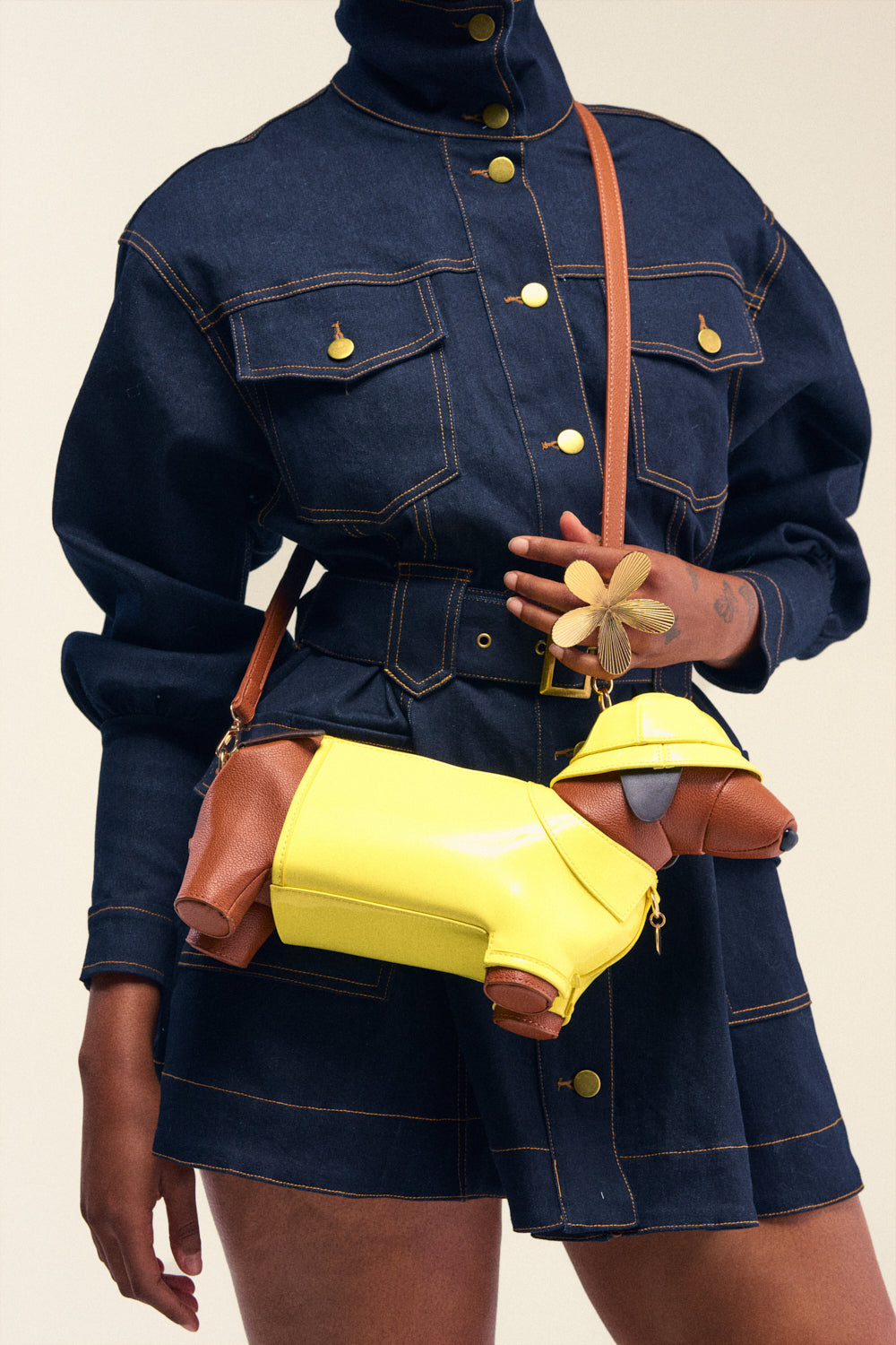 A person in a dark denim outfit holds the WOOF PURSE—a brown and yellow dachshund-shaped novelty handbag with a yellow hat—worn across the body with a crossbody strap. They also have a flower-shaped accessory on their hand.