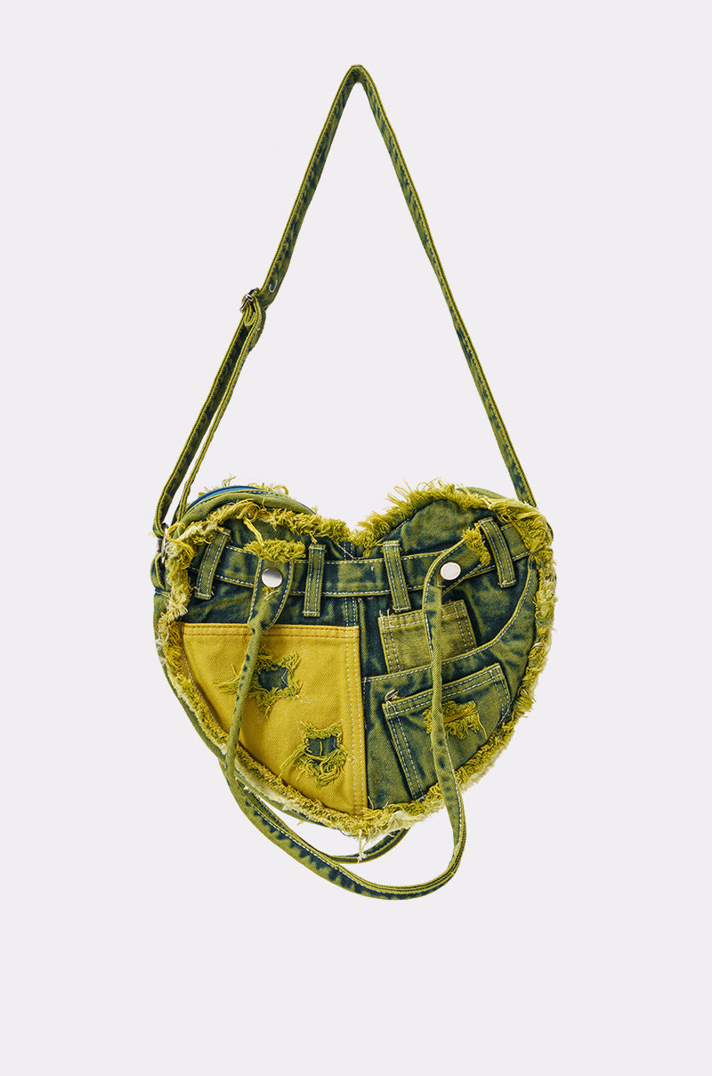 The WHATYA WANT DENIM PURSE features green and yellow heart-shaped denim patches, visible stitching, frayed edges, multiple pockets, silver rivets, and an adjustable strap for a unique upcycled festival look.