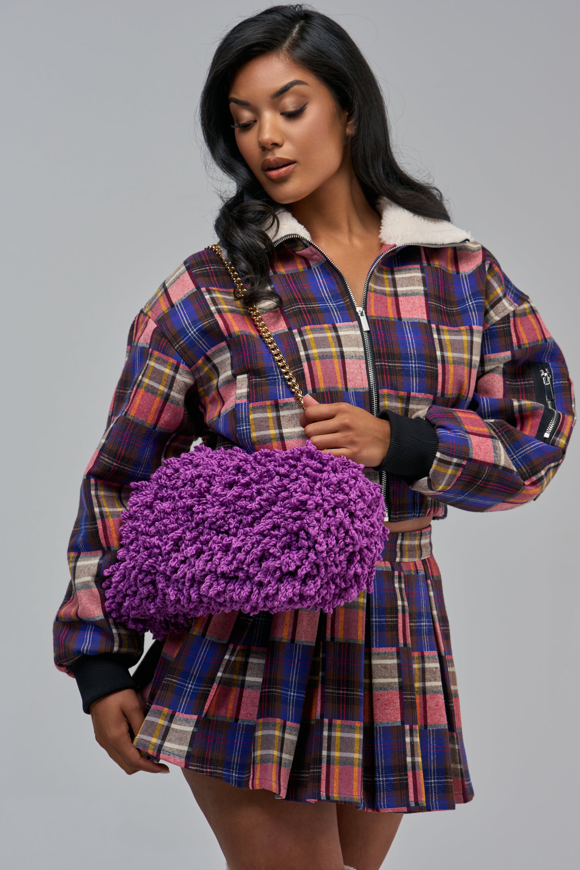 A person wearing a plaid jacket and skirt holds an IT IS WHAT IT IS CLUTCH, a purple, shaggy textured baguette purse with a gold chain strap.