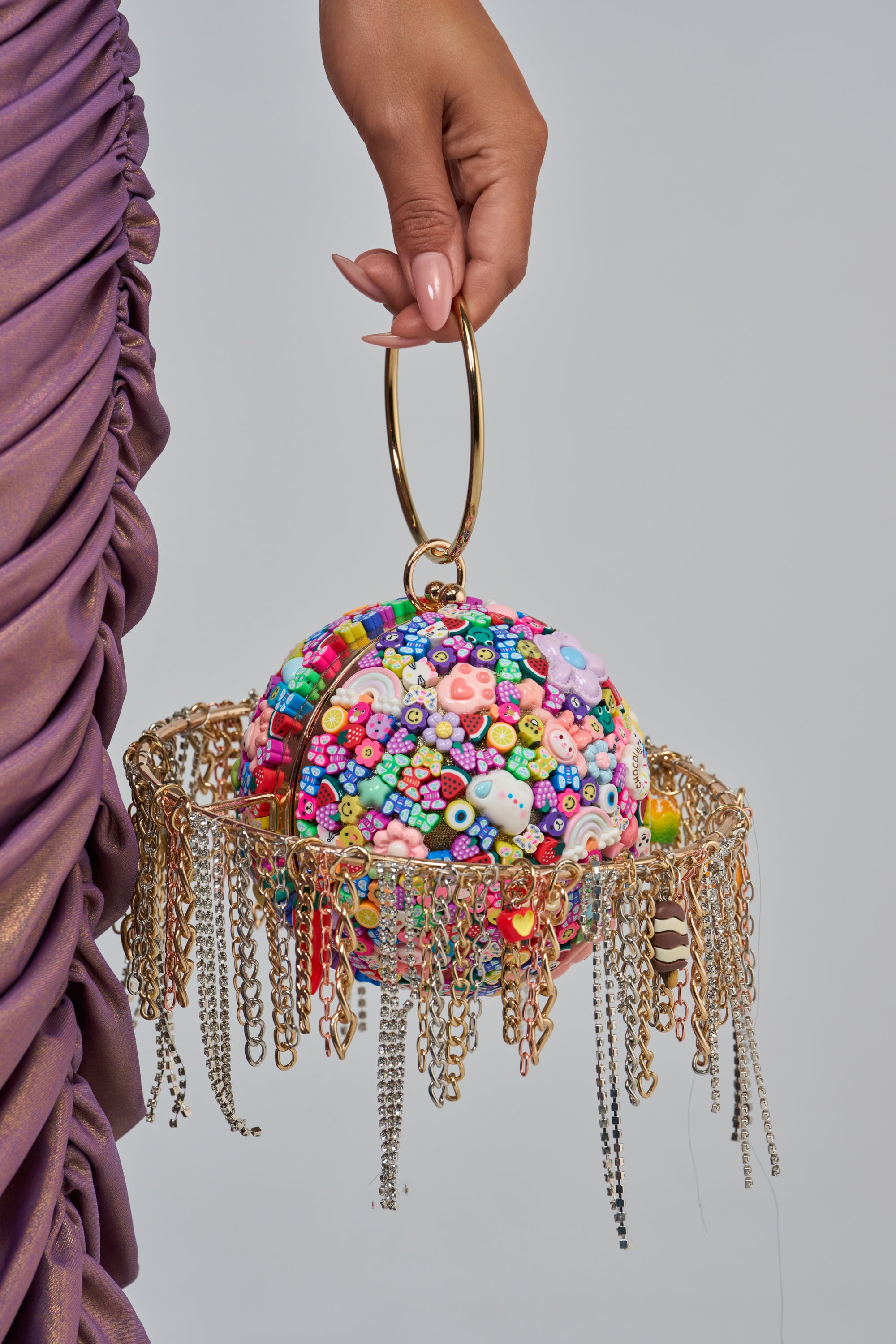 A hand holds the OUT OF THIS WORLD NOVELTY PURSE, a round statement bag adorned with colorful beads, gold rings, and dangling chains, next to a ruched purple dress.