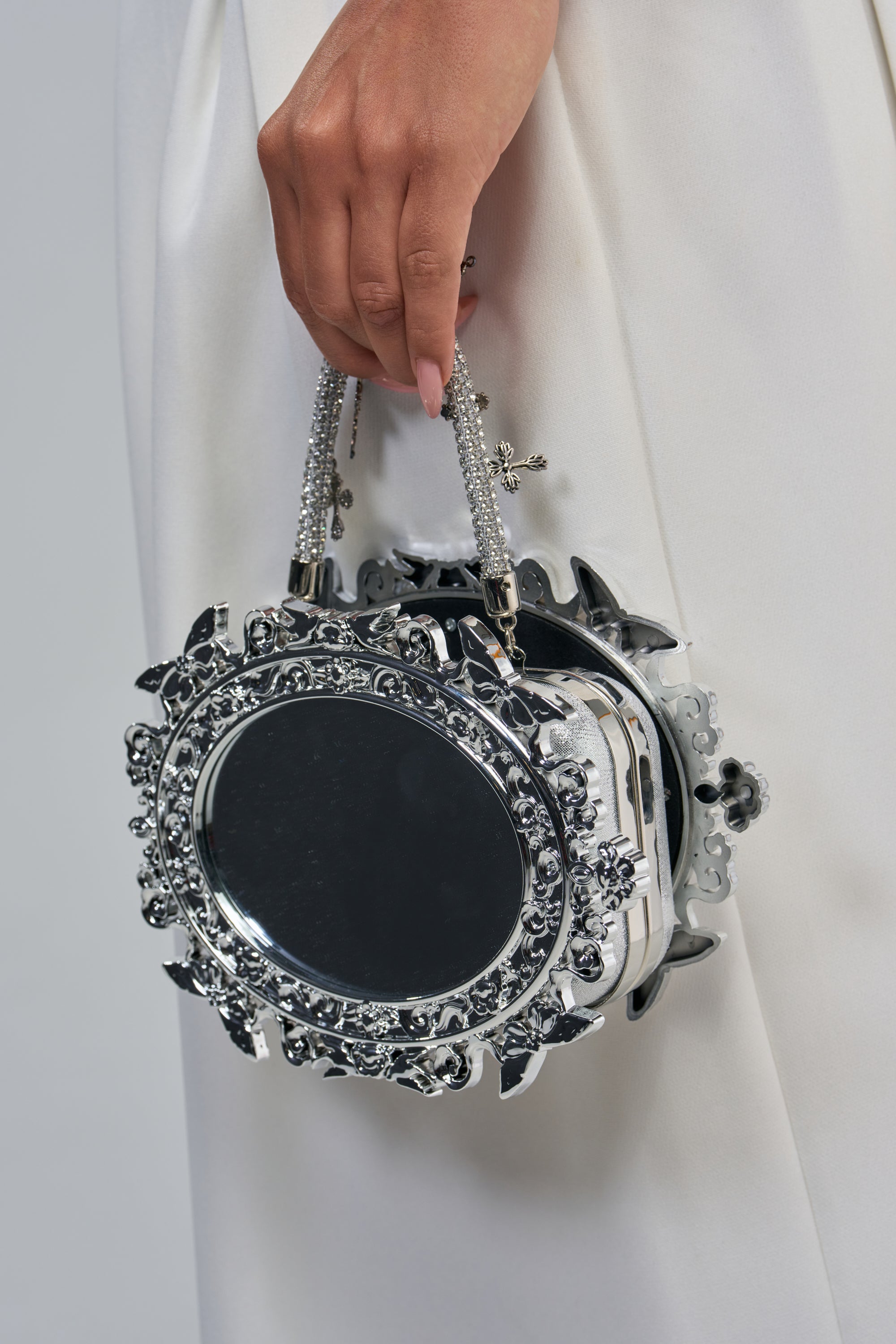 A hand holding the LOOK AT ME MIRROR BAG—a round, baroque-inspired silver handbag adorned with decorative jewels and a mirrored center—makes for the ultimate statement accessory against a white outfit background.