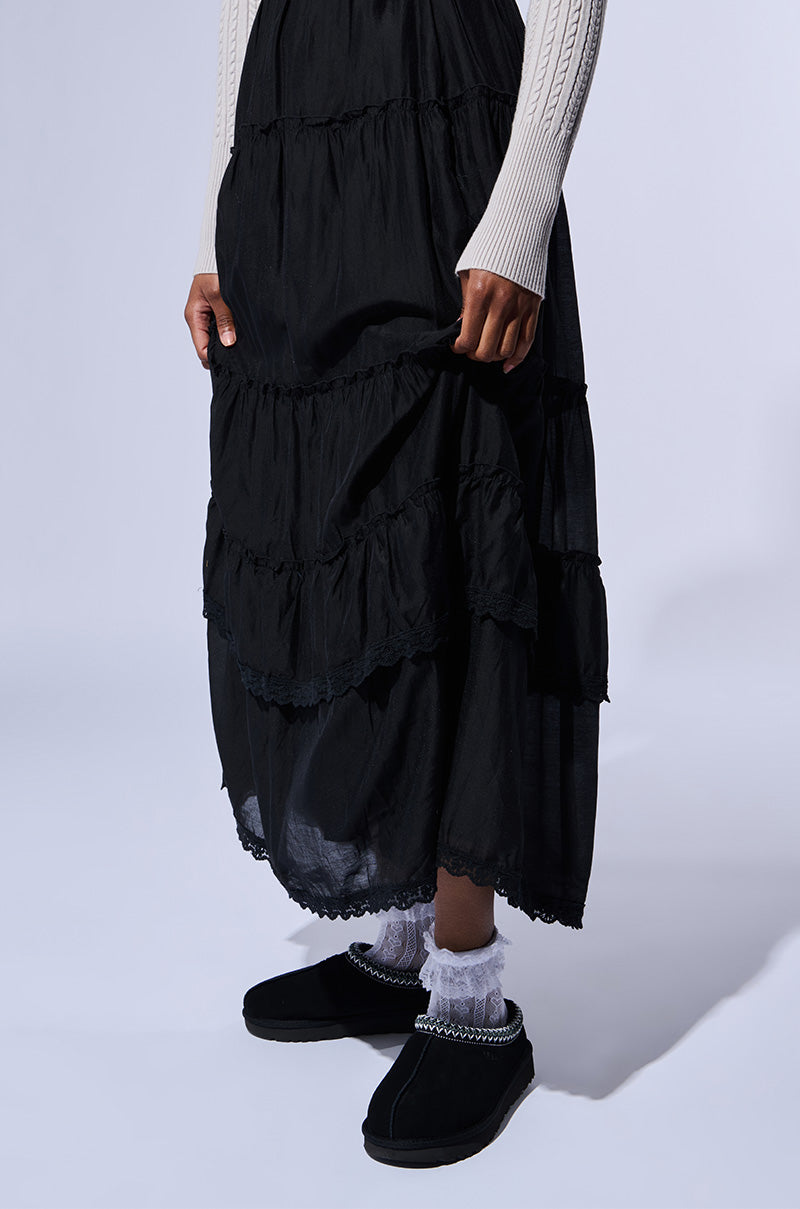 A person in a black tiered skirt and cream ribbed sweater stands against a plain background, wearing the UGG Tasman Slipper in Black, which adds an elegant touch with its suede upper and pearl detailing. White frilly socks are visible as they hold the skirt with one hand.