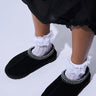 A person wearing UGG Tasman Slippers in black, featuring suede uppers with intricate stitching, paired with white lace socks. The person is dressed in a black skirt with a lacy hem, standing on a plain white surface.