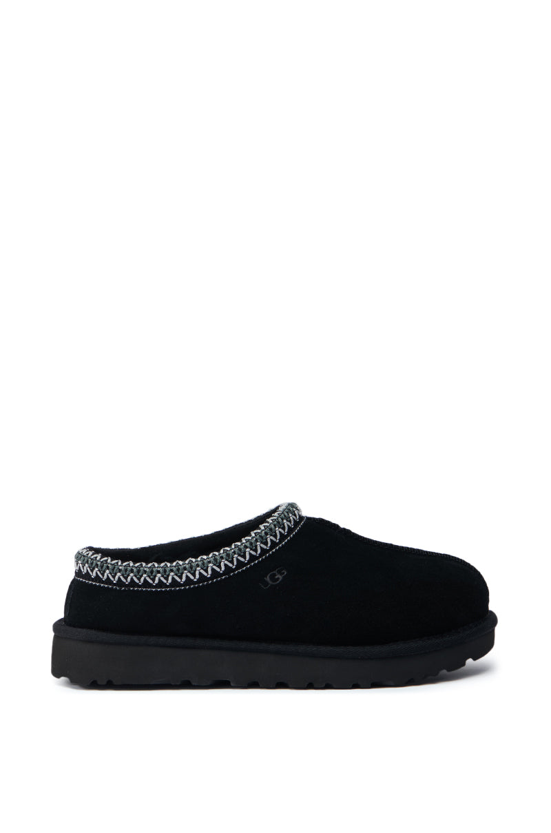 The UGG TASMAN SLIPPER IN BLACK boasts a soft, rounded design with decorative stitching around the ankle. It incorporates an upcycled wool lining and a flat, textured sole for enhanced grip. With its suede upper adding a touch of elegance, this slipper seamlessly combines style and sustainability.