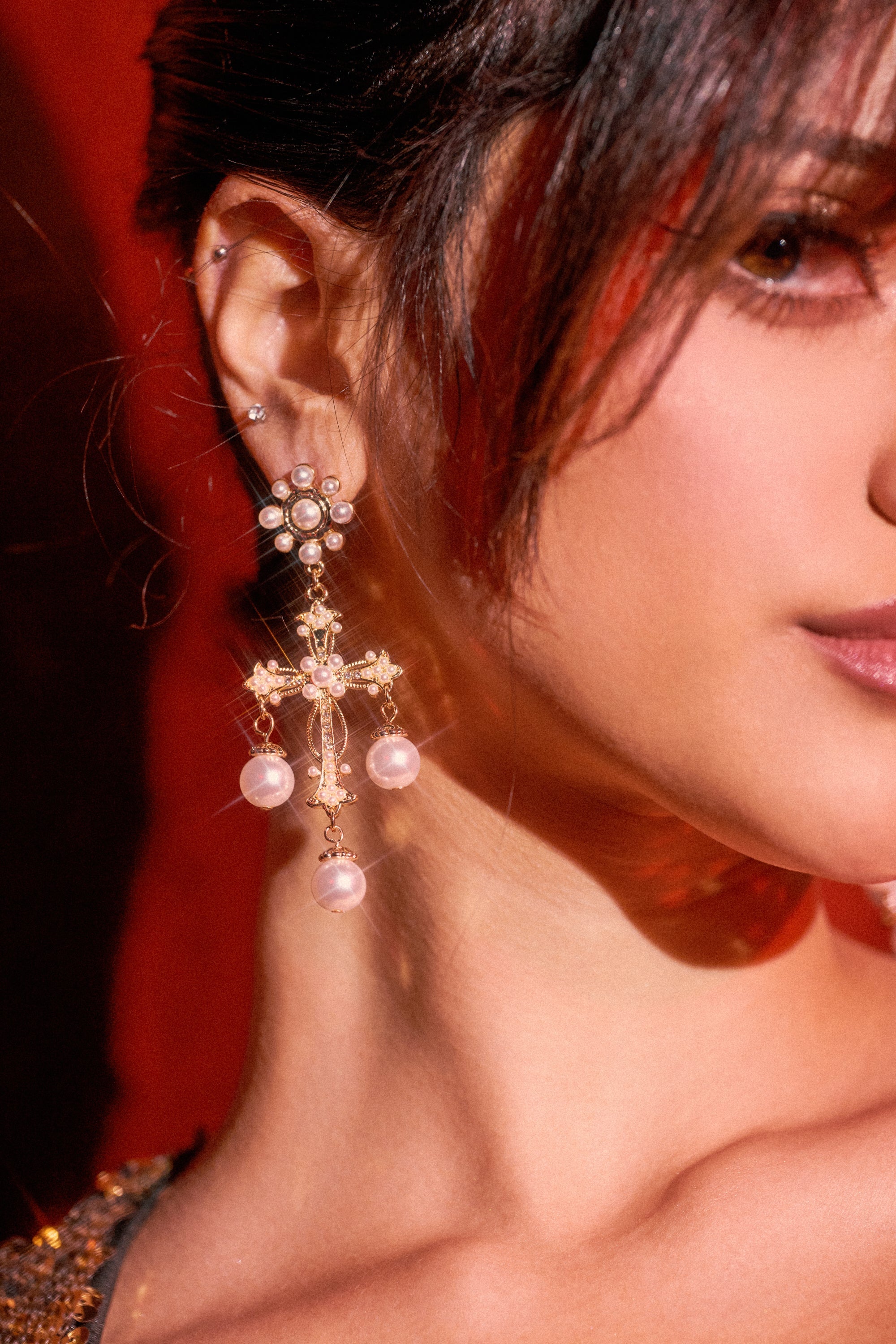 Close-up of a woman's ear wearing the CROSS IT OUT EARRING, a large ornate cross design with pearls. Part of her face and dark hair appear against a red background. 0