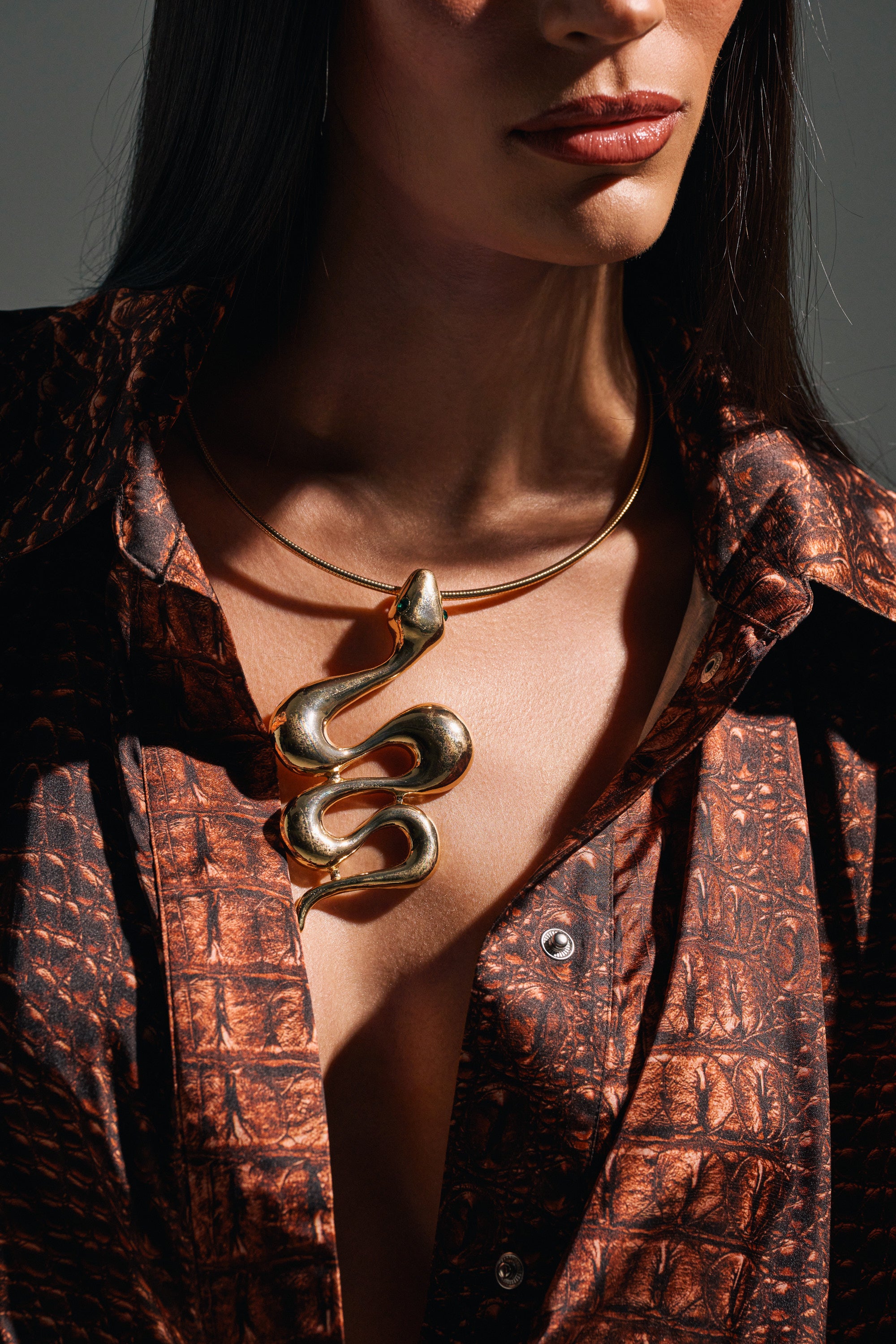 A woman in a textured brown-patterned shirt wears the WATCH OUT FOR THEM NECKLACE, a bold gold snake pendant. Her face is partially visible, drawing attention to her lips and dark hair as she showcases the statement piece.