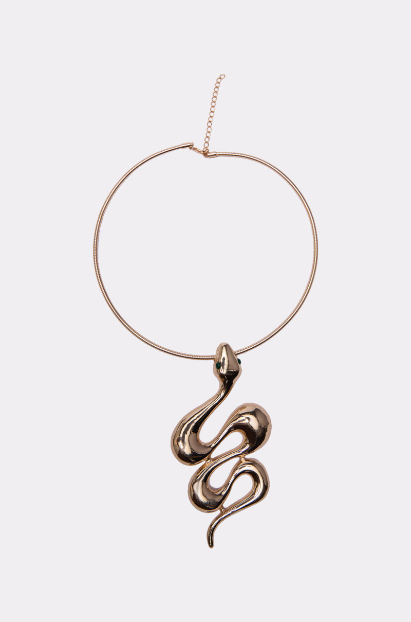 The WATCH OUT FOR THEM NECKLACE is a gold choker featuring a bold, abstract snake-shaped pendant with a vivid green gemstone, shown against a simple light background.