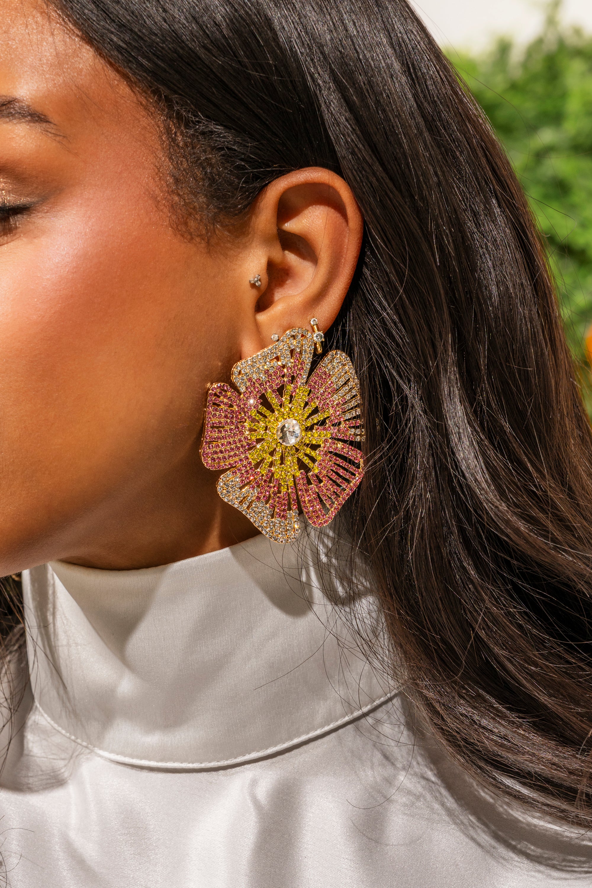 A close-up of a woman with long brown hair in a white high-collared top, wearing the IM JUST PRETTY EARRING—bold floral rhinestone earrings set with yellow, pink, and clear crystals. 0