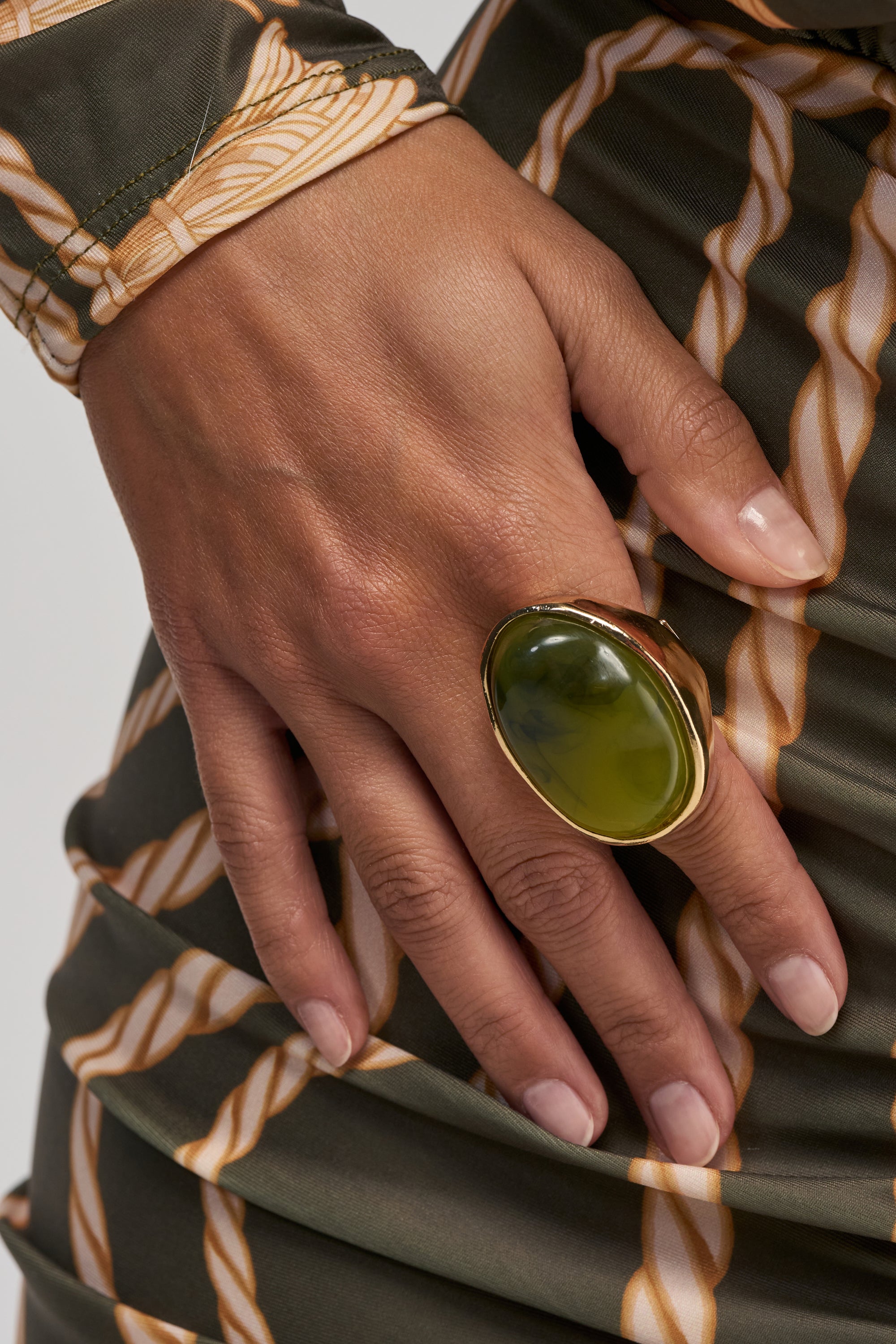 A hand displays the VACATION NIGHTS GREEN STONE RING, featuring a large oval green gemstone set in gold, posed against olive green and gold printed fabric—an eye-catching statement stretch ring ideal for boho-chic jewelry lovers.