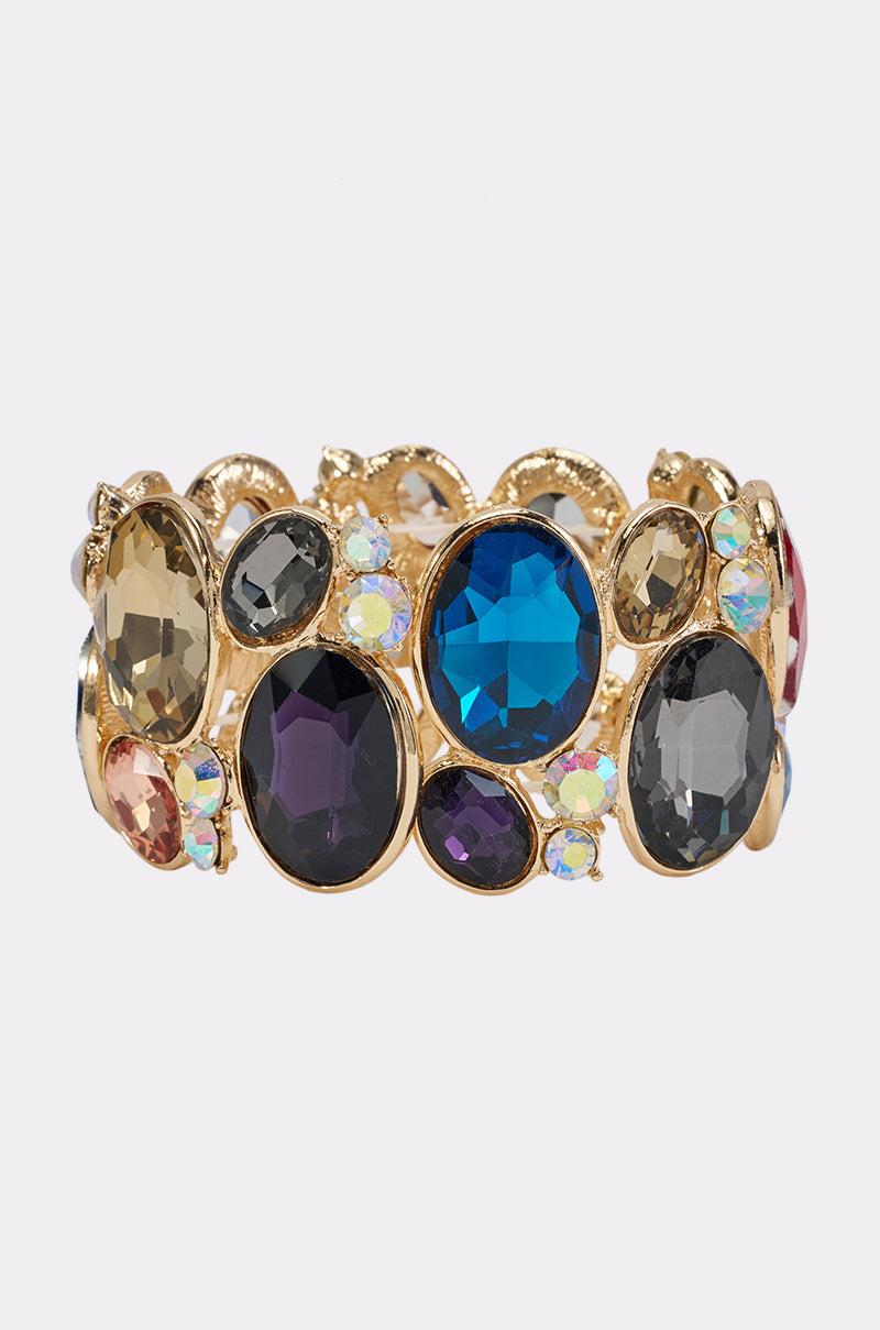 The MULTI-STONE STRETCH BRACELET features an ornate gold design embellished with large, multi-colored gemstones in shades of blue, purple, black, and iridescent hues—making it a striking statement accessory with its intricate crystal details.