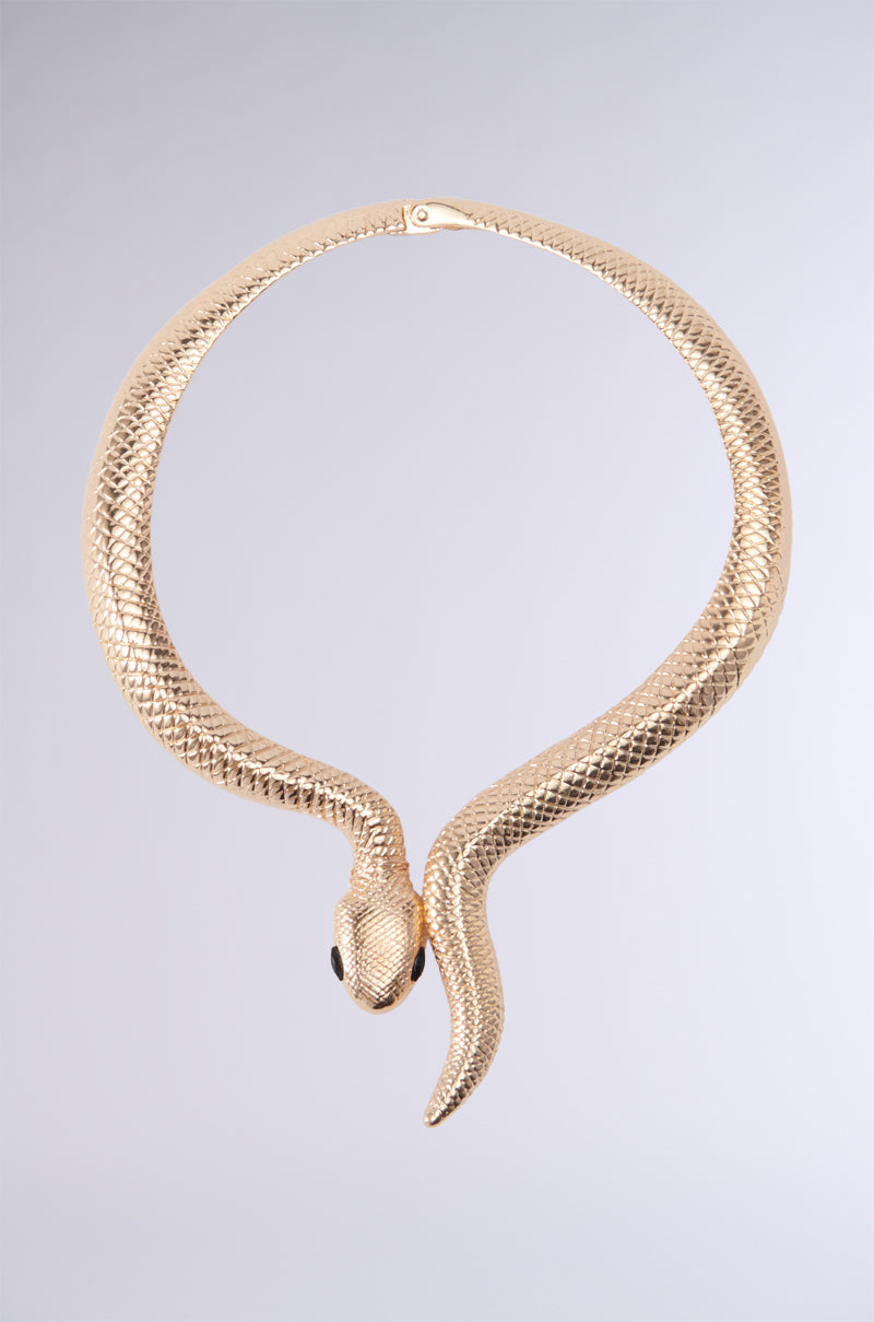 The BITE ME NECKLACE is a striking gold piece designed to resemble a coiled snake with an intricate textured pattern. It features two small, black gemstone eyes and has its head and tail meet at the front center, creating an elegant and unique statement jewelry piece. This adjustable necklace stands out vividly against a plain gradient gray background.