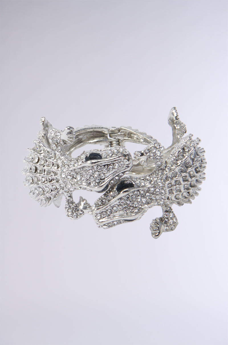 The REPTILE MANIA CUFF is an ornate faux silver metal bracelet that showcases the intricate designs of two rhinestone-embellished reptiles entwined together. Detailed with numerous small, sparkling crystals, the alligators face each other and form a circular shape against a plain light gray background. 0