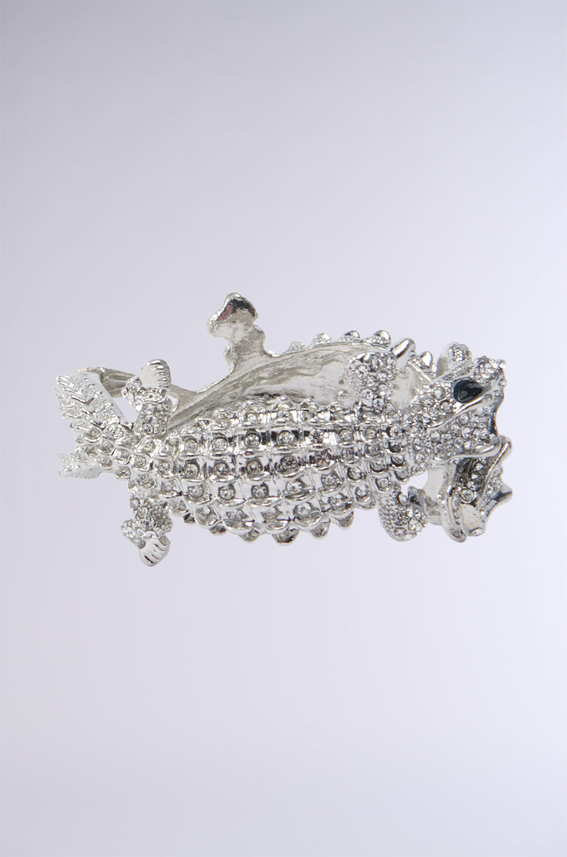 The REPTILE MANIA CUFF is a silver, textured accessory depicting a stylized, abstract creature with a rough, craggy surface and intricate details, resembling rhinestone-embellished reptiles. Its metallic finish reflects light, highlighting its elaborate craftsmanship. 2