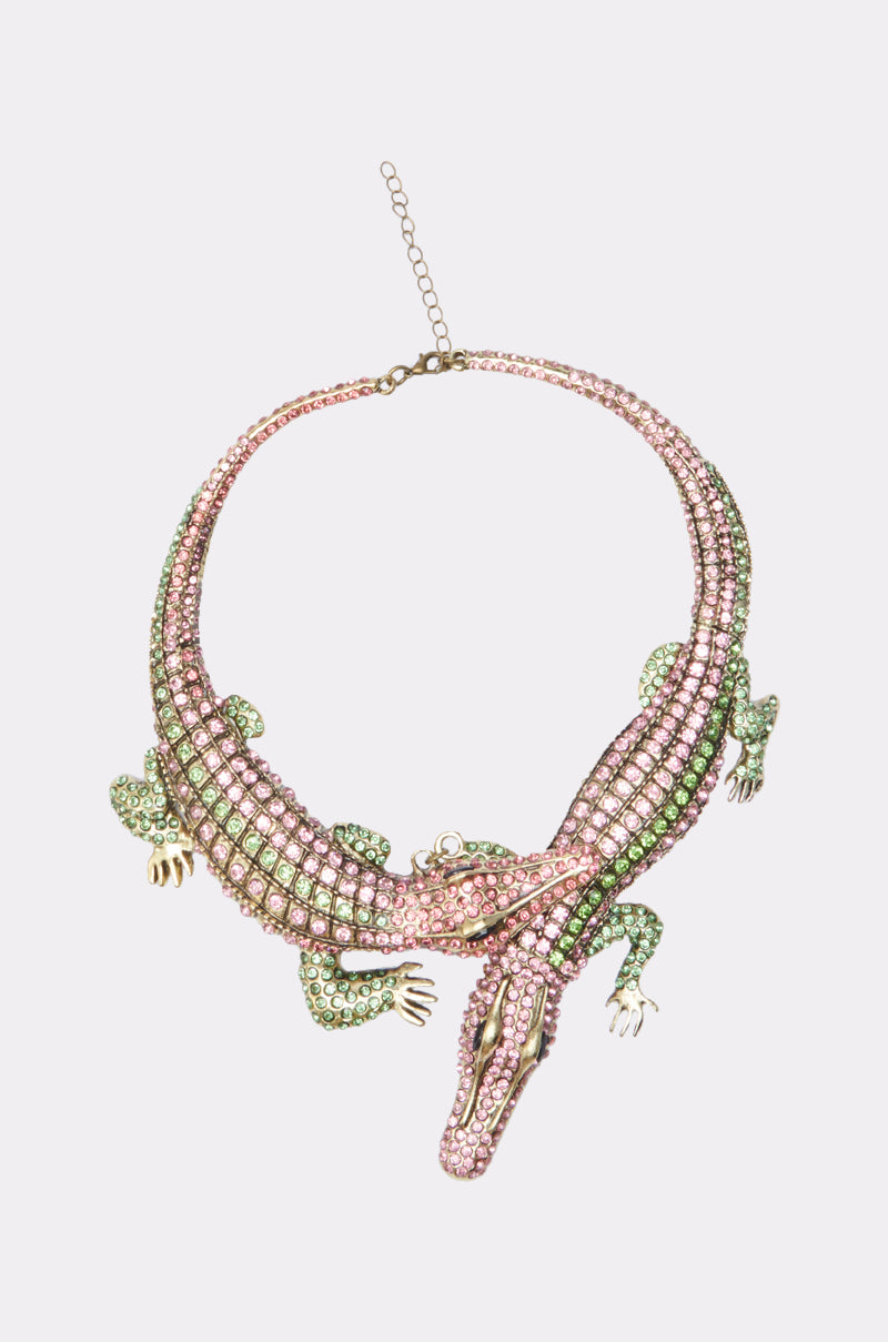The I BITE BACK NECKLACE is a bold statement piece with two interlocking alligators adorned in pink and green rhinestones, gold accents, and an adjustable chain clasp.