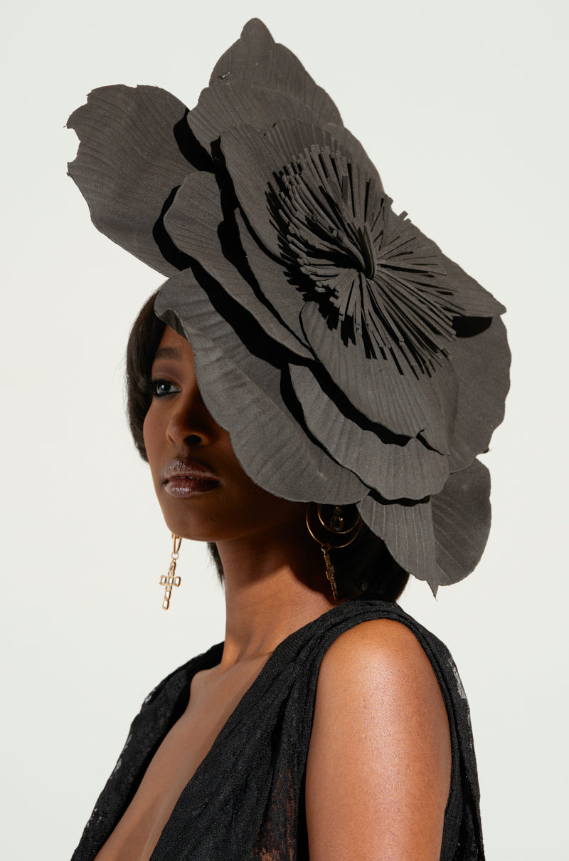 A woman wears the IN BLOOM HAIR ACCESSORY, an elegant large black floral fascinator with layered petals. It beautifully complements her black outfit and gold cross-design earrings, standing out against the plain, soft light background.
