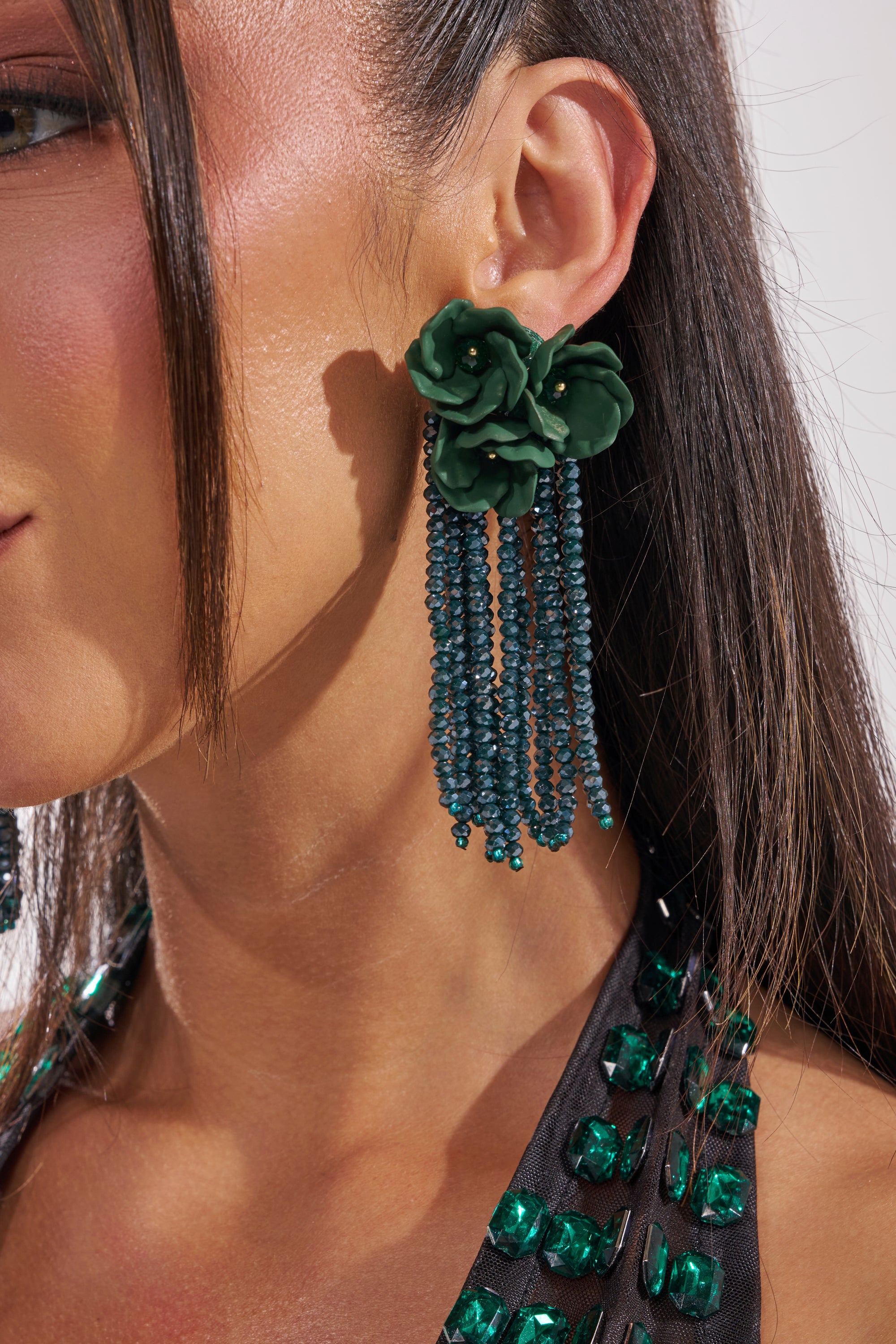 A close-up of a woman's ear wearing the BERLIN EARRING, featuring a green floral drop design with beaded fringe strands. Her straight hair is tucked back, revealing a top embellished with green gems. 1