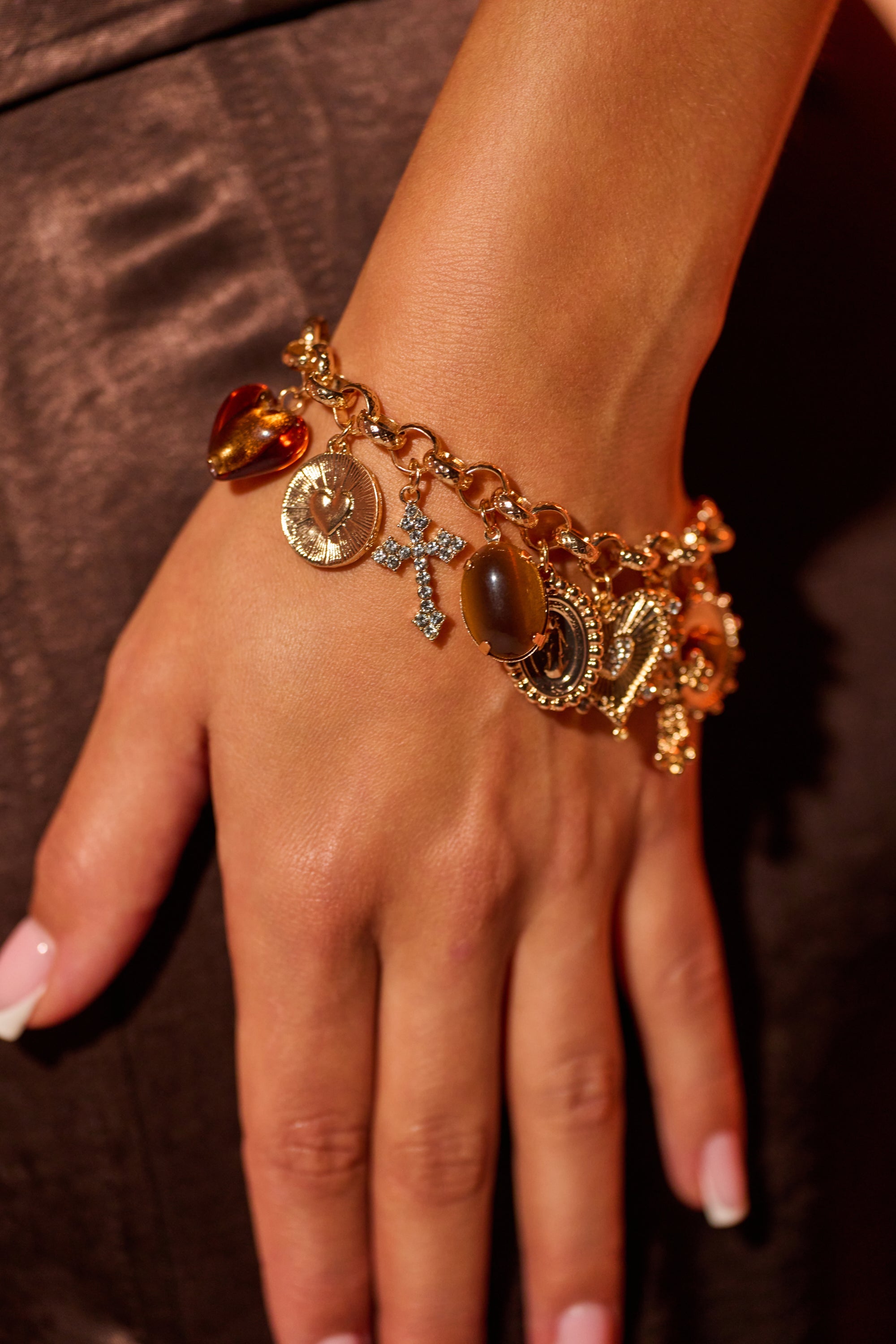 A hand with manicured light pink nails wears the CHARM IT UP BRACELET, featuring a gold-tone chunky link chain and vintage-inspired cross, heart, and circular charms, resting elegantly against brown satin fabric. 0
