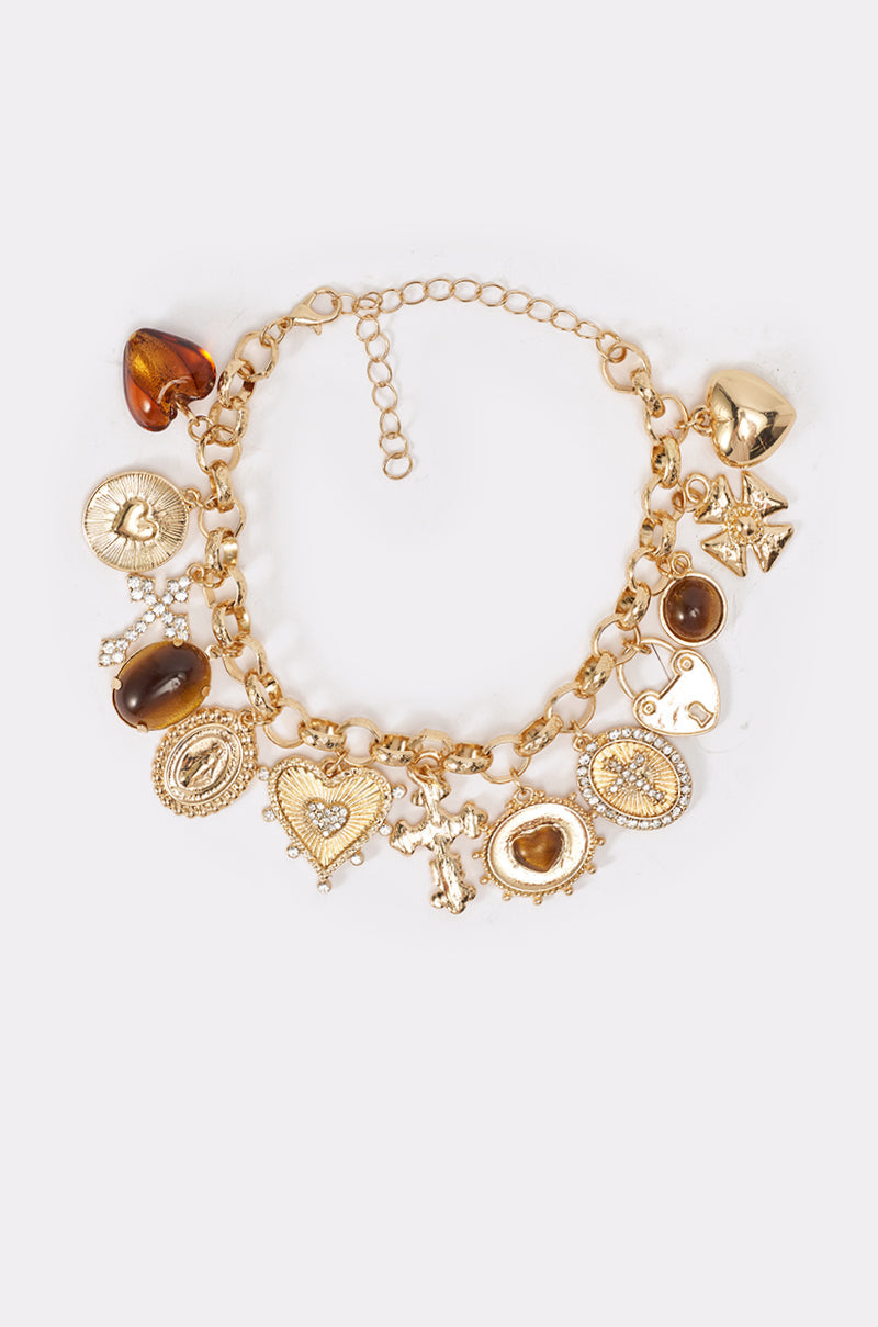 The CHARM IT UP BRACELET is a gold-tone charm bracelet with vintage-inspired hearts, crosses, coins, stars, and amber-like stones, shown on a light background. 2