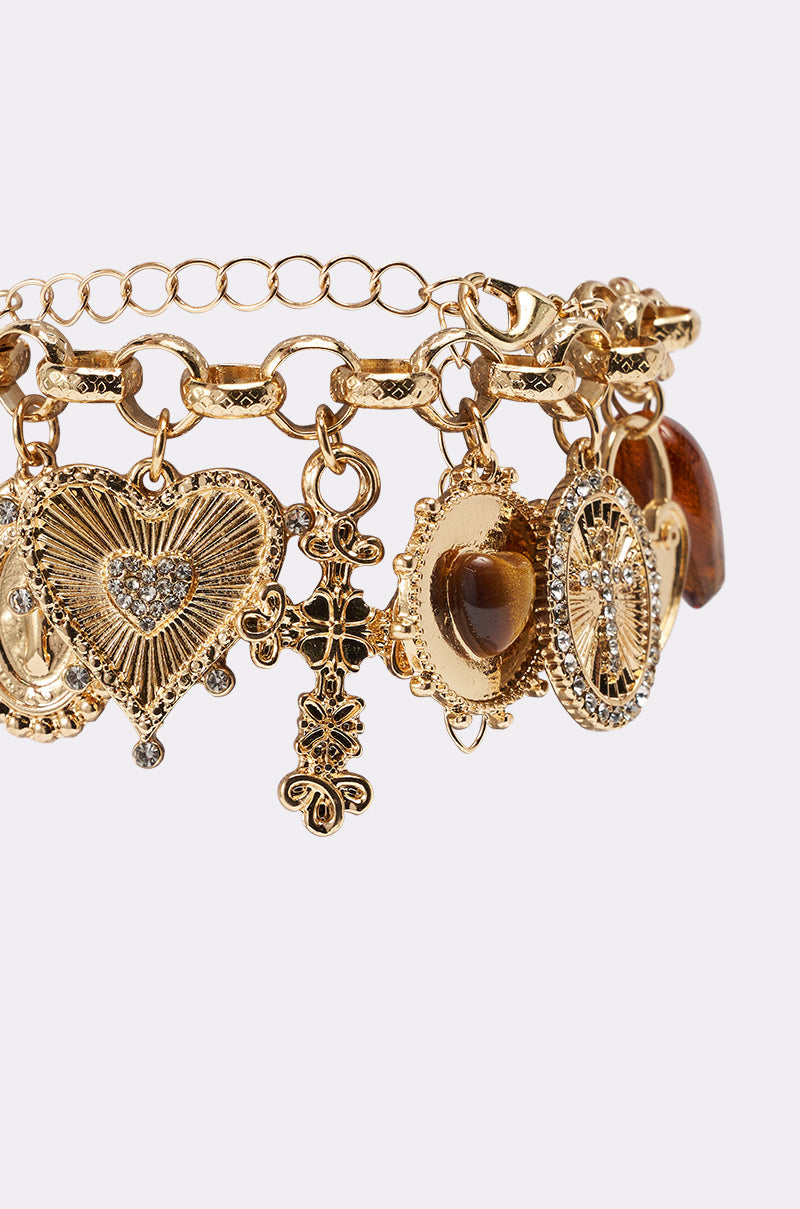 The CHARM IT UP BRACELET features a chunky gold-tone link chain with vintage-style heart, cross, oval, and round charms—some decorated with rhinestones and brown gemstone accents—shown on a white background. 3