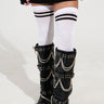 A person stands wearing knee-high, black gothic boots embellished with chains and buckles, paired with the ULTIMATE THIGH HIGH SOCKS in white featuring varsity stripes. The outfit is completed with a black skirt and long sleeves.