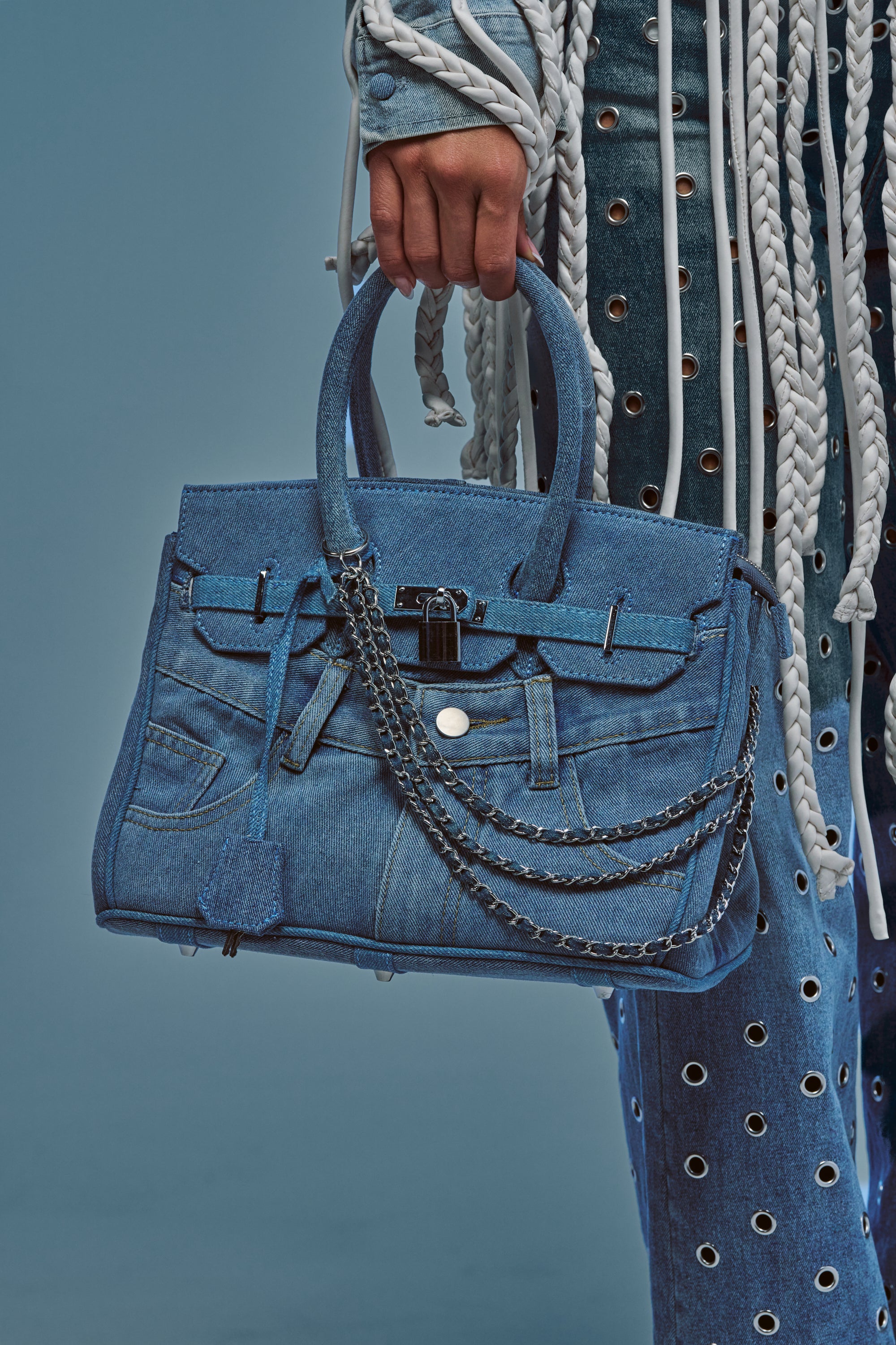 A person holds the DENIM BLUES TOTE PURSE, a blue denim tote bag with chain details, while wearing coordinating streetwear-inspired denim clothing adorned with grommets and white braided cords, set against a blue background.