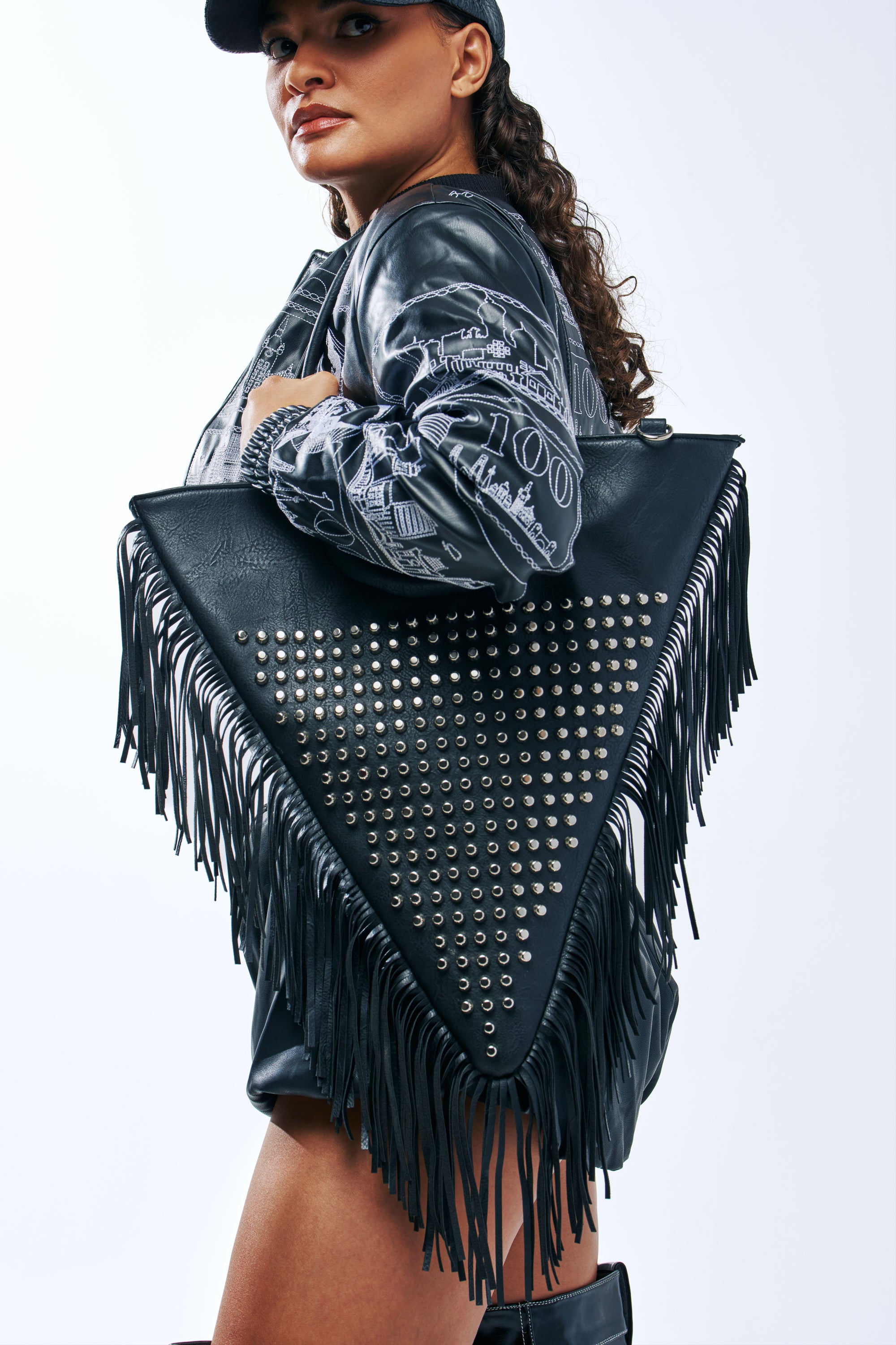 A model wearing a black jacket carries a large, triangular TAKE ME OUT STUDDED TOTE with fringe.