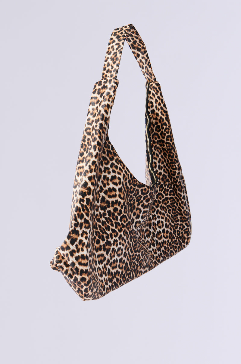 The WILD ONE PU OVERSIZED TOTE features a chic, slouchy design with a leopard print of brown and black spots on a light background. The tote has a wide strap and faux leather material, displayed against a plain gray backdrop for an effortlessly striking look suitable for any occasion.