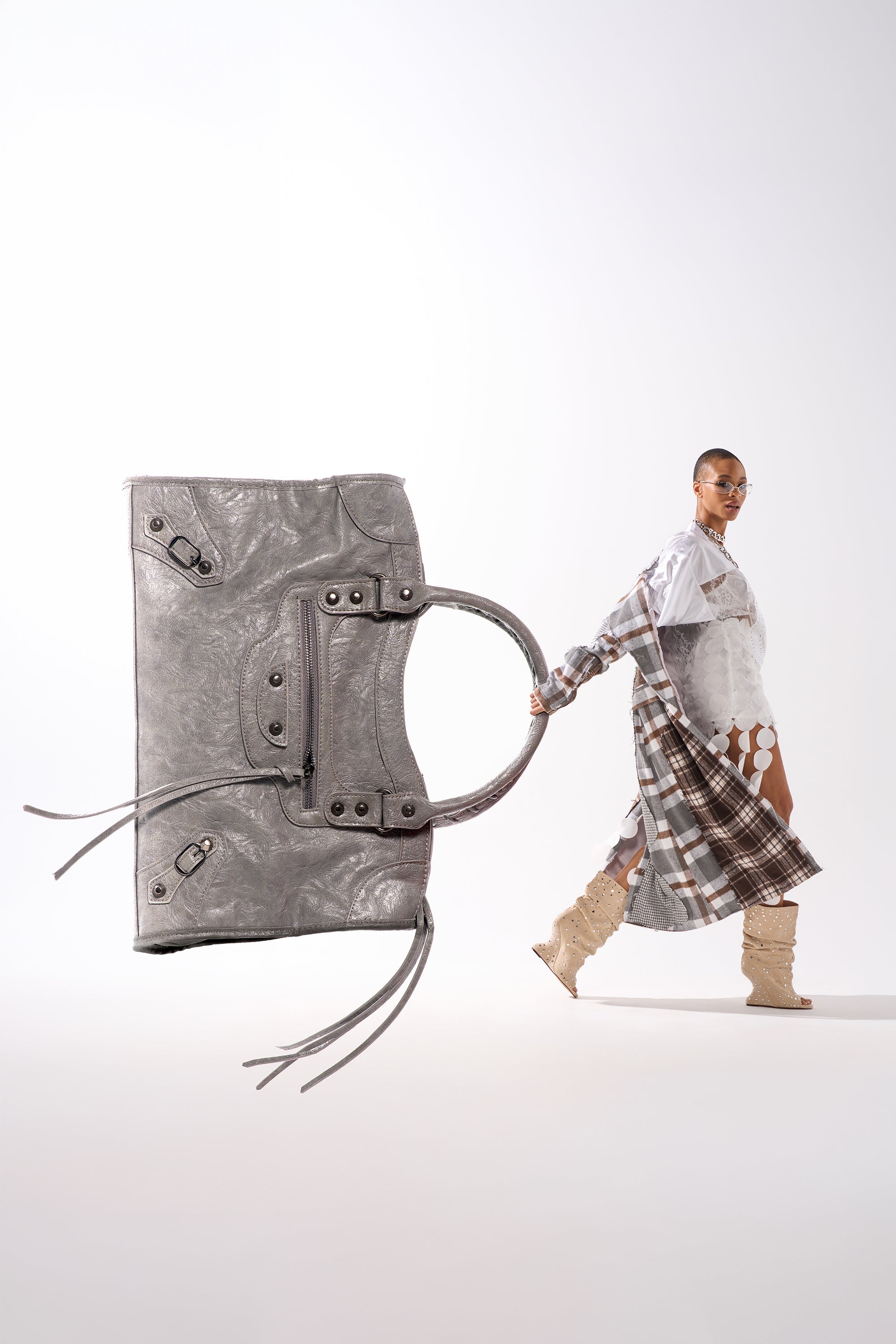 The ROSIE OVERSIZED PU TOTE in silver is featured prominently, with a model in a patterned dress and beige boots walking confidently in the background against a plain white backdrop.