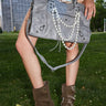 A person stands on grass wearing brown boots and a beige animal-print skirt, holding the ROSIE OVERSIZED PU TOTE with pearl chains. Tall buildings and green trees are visible in the background.