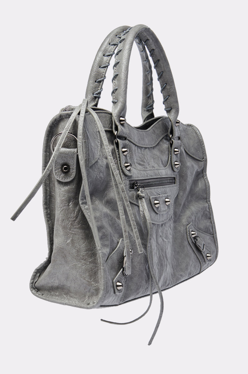 The ROSIE OVERSIZED PU TOTE is a gray faux leather handbag with dual handles, silver studs, a front zip pocket, and long tassels on the zippers, pictured at a three-quarter angle against a light background.