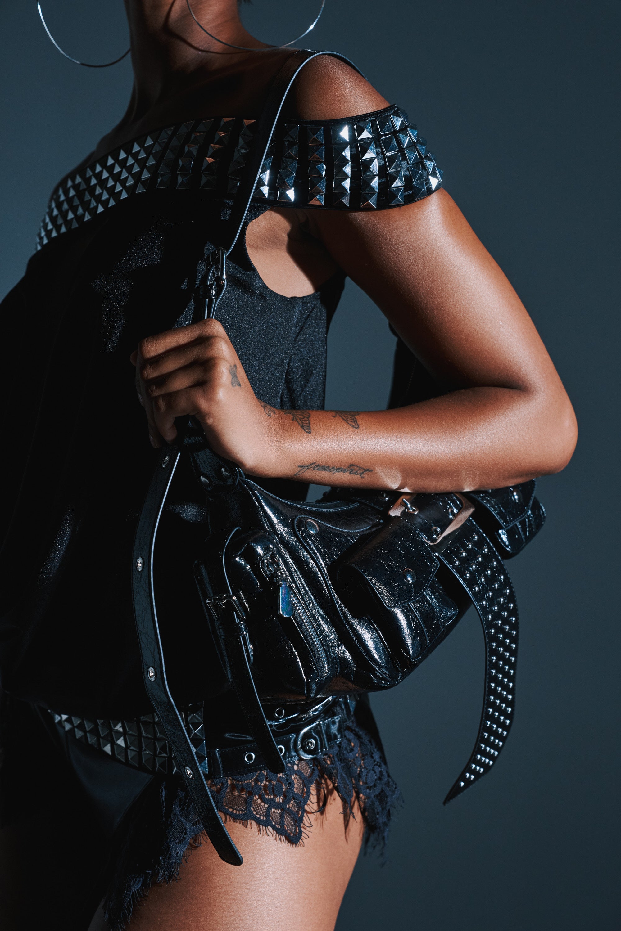 Wearing a studded black top and lace shorts, a person holds THE TIDUS PU PURSE— a faux patent leather bag—against a dark background. Only their lower face and upper body are visible. 1