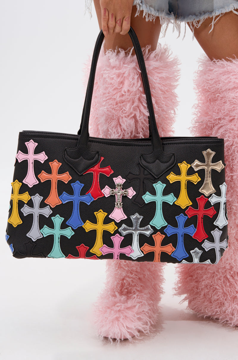 A person in pink furry boots and frayed denim shorts carries the PERSONAL PROBLEM CROSS TOTE, a large faux leather bag adorned with vibrant cross-shaped patches. 1