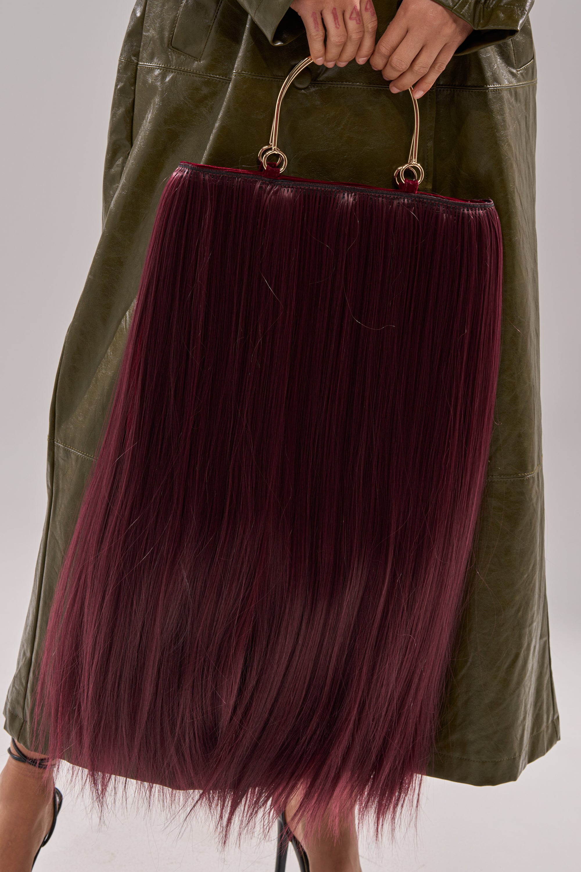 A person in an olive green coat holds the MY HAIR PURSE, a large handbag with long, dark burgundy hair-like strands and a gold-tone handle, making for an eye-catching and unusual accessory.