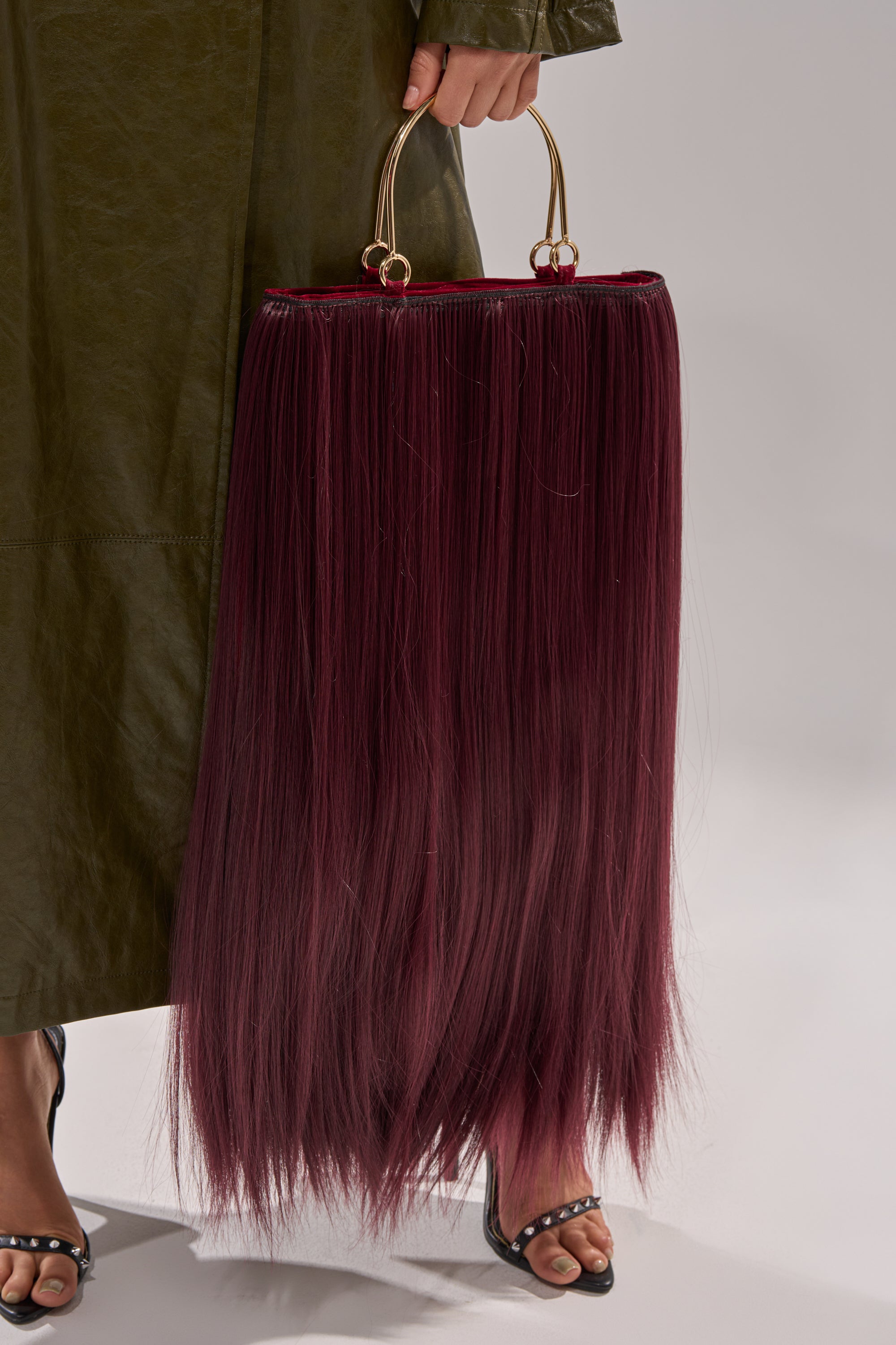 A person holds the MY HAIR PURSE, featuring long, straight burgundy hair-like strands and gold handles, paired with a dark green coat and black sandals.