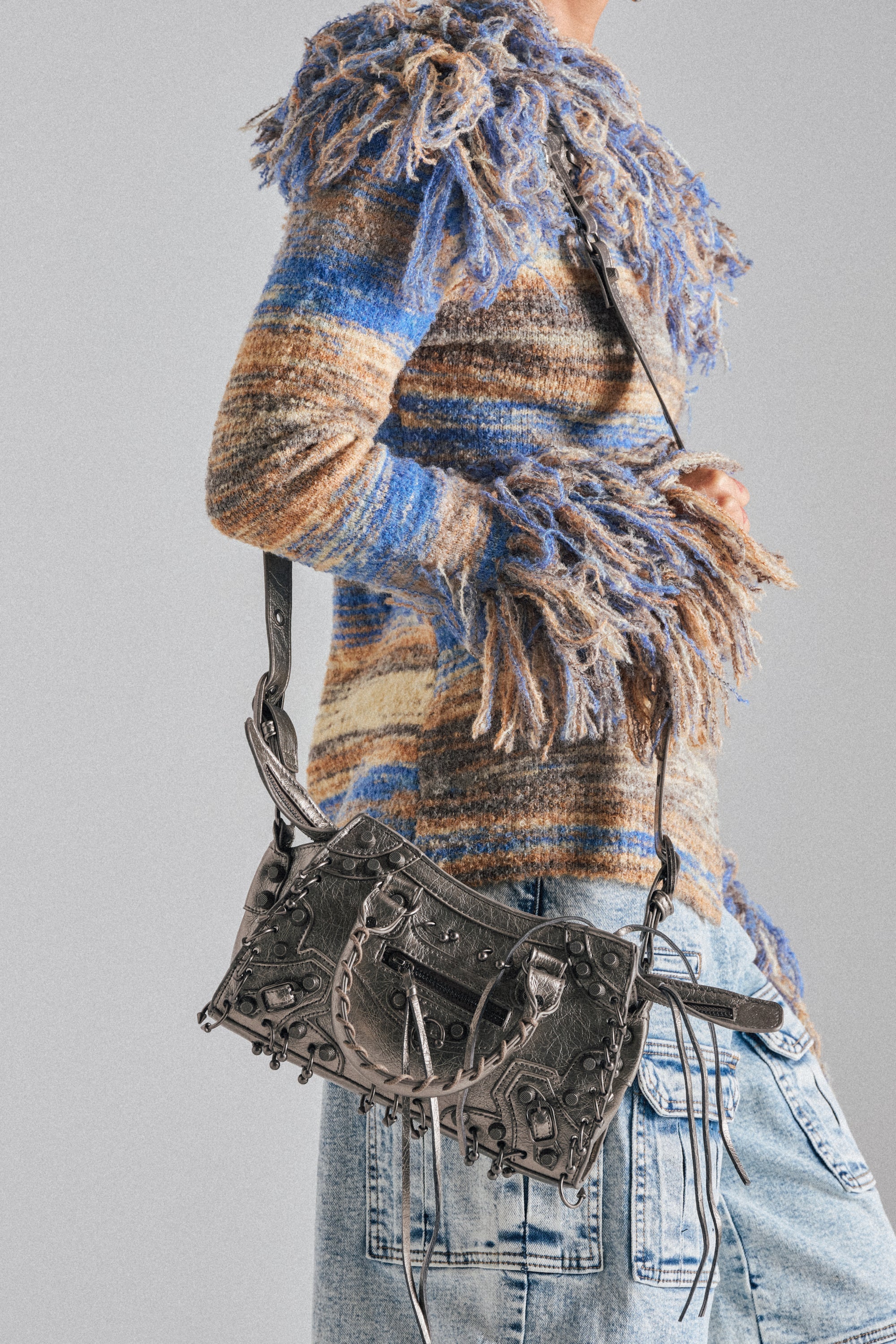 A person in a fuzzy, multicolored striped sweater and distressed jeans carries the GRUNGE PIERCED PURSE, a textured faux leather handbag with tassel details. The background is plain gray. 1