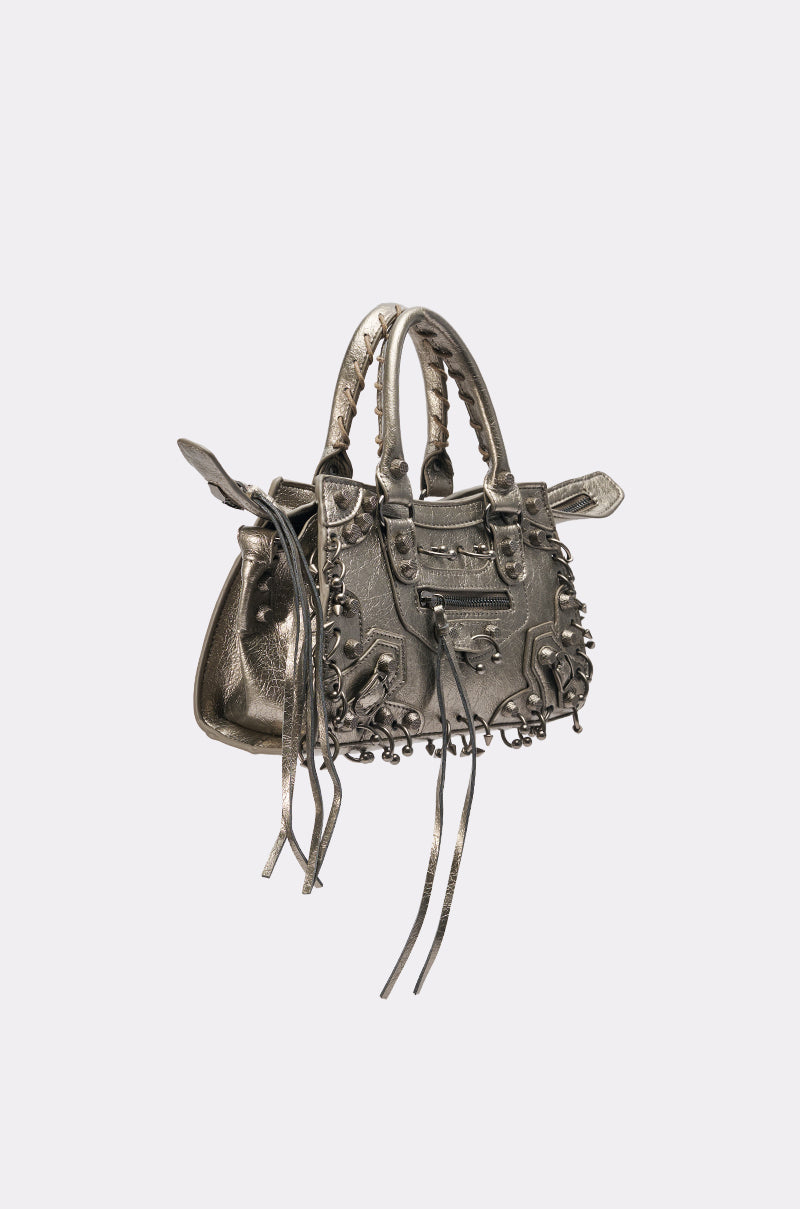 The GRUNGE PIERCED PURSE is a metallic silver handbag featuring braided handles, pierced hardware, decorative studs, and long tassels on the zippers, shown against a plain white background.