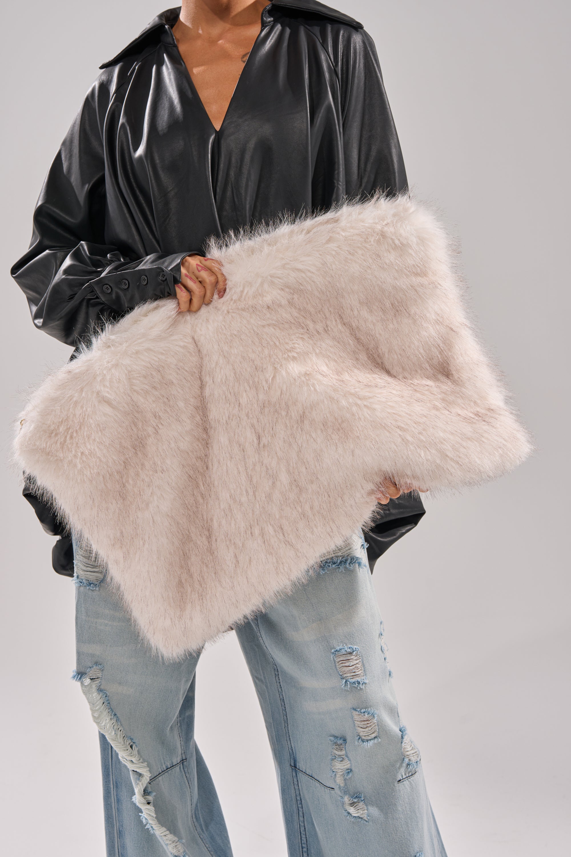 A person holds the CRU FAUX FUR OVERSIZED CLUTCH, a winter essential, while wearing a black leather jacket and ripped light blue jeans against a plain background. 0