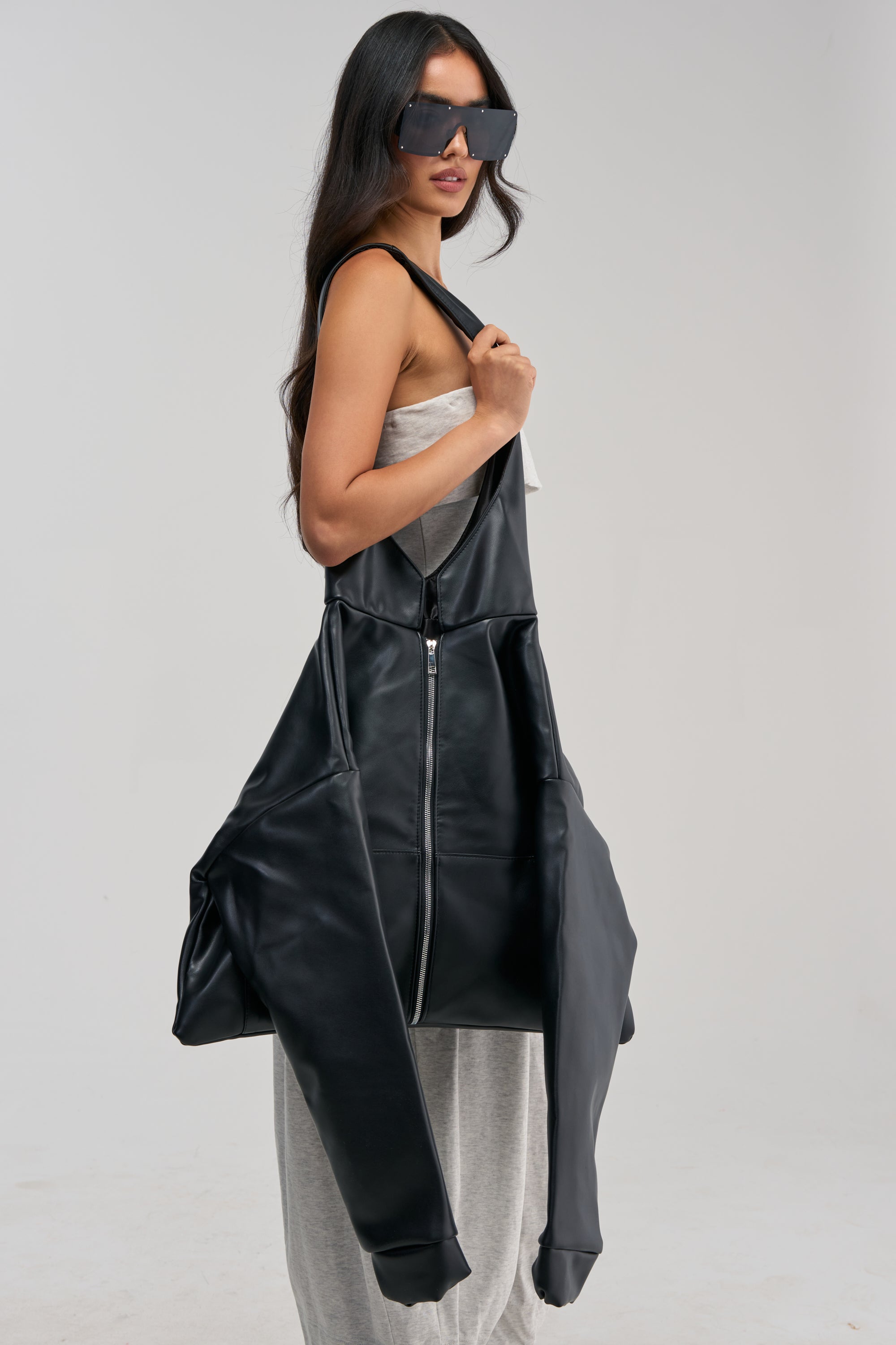 A woman in a gray dress and large sunglasses carries the PUT YA PANTS ON PURSE, an oversized faux leather bag styled to resemble a jacket with sleeves and a zipper, set against a plain light background.