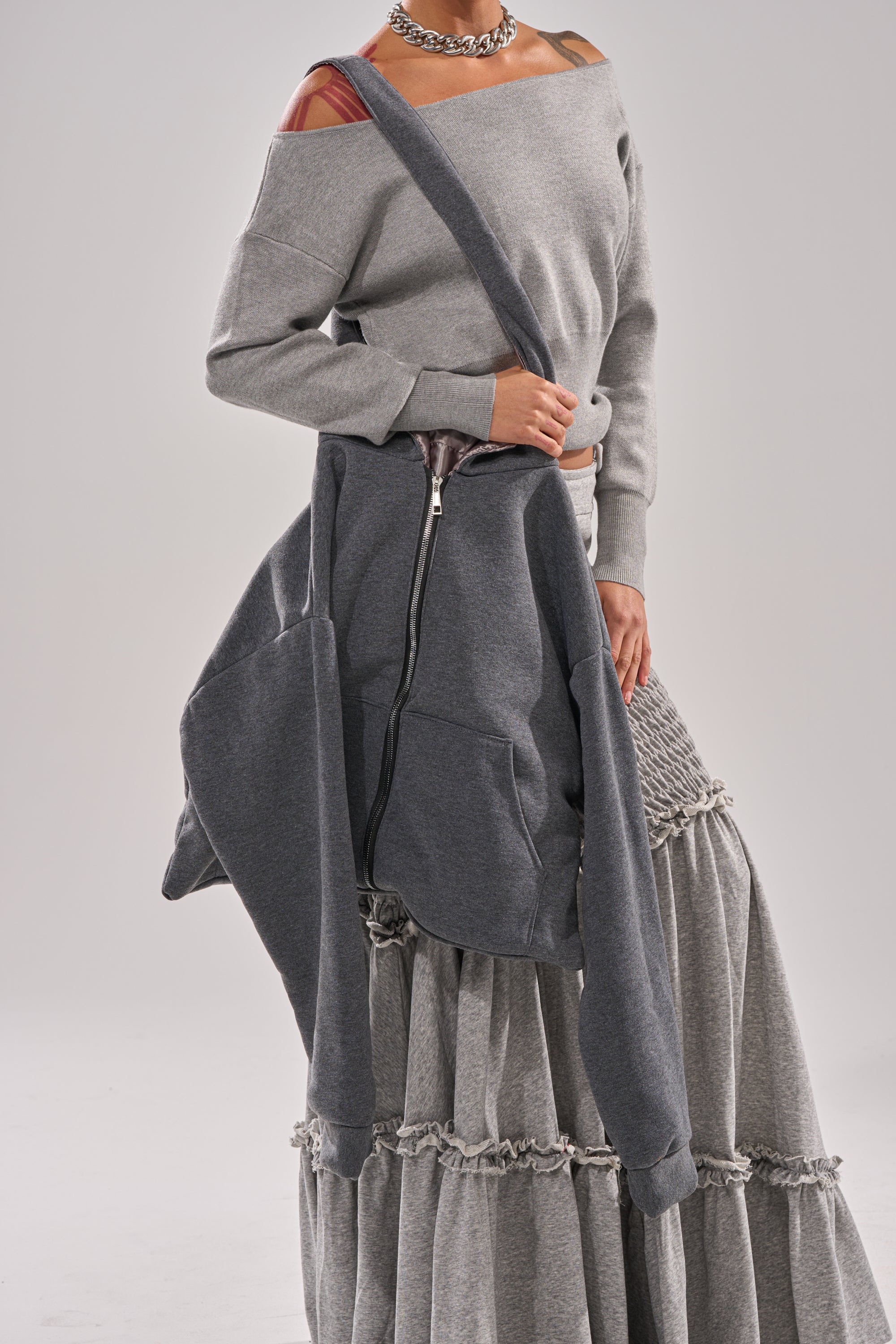A person, visible from lower face to torso, sports an off-shoulder gray sweater and layered ruffled skirt while carrying the CARRY ME HOODIE PURSE—a large, street-inspired gray accessory slung across the body for an effortlessly cool look.