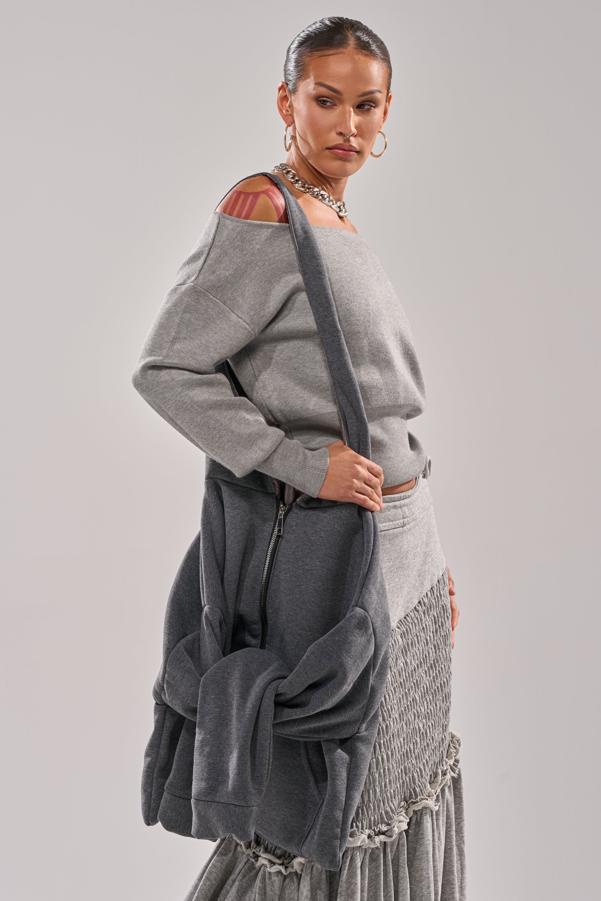 A woman stands against a neutral background wearing a stylish off-shoulder grey top, textured skirt, hoop earrings, chunky chain necklace, and holding the CARRY ME HOODIE PURSE. She looks down with a serious expression.