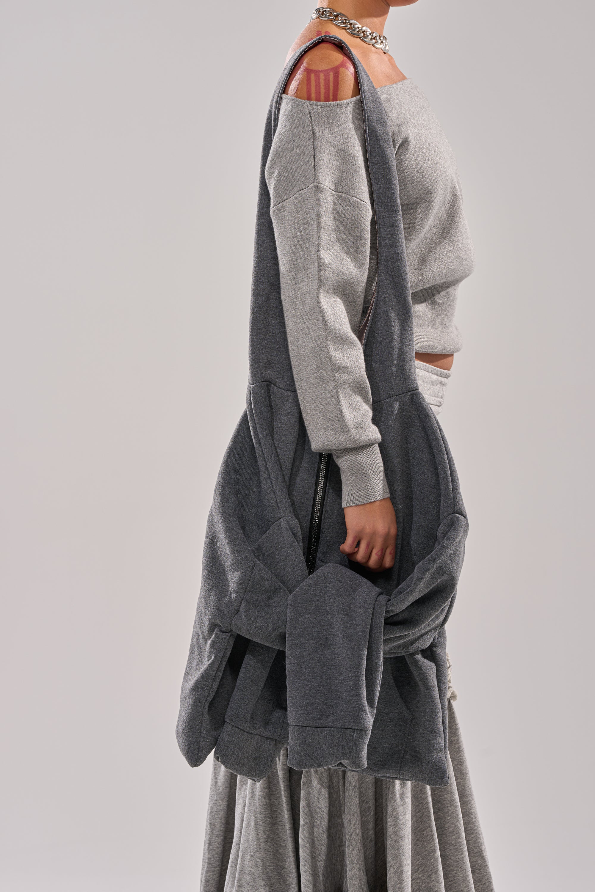 Wearing a gray off-shoulder sweatshirt that reveals a pink-striped bra strap, the person carries the CARRY ME HOODIE PURSE—a street-inspired, large gray bundled cloth bag—and pairs it with a matching skirt. The background is plain and light.