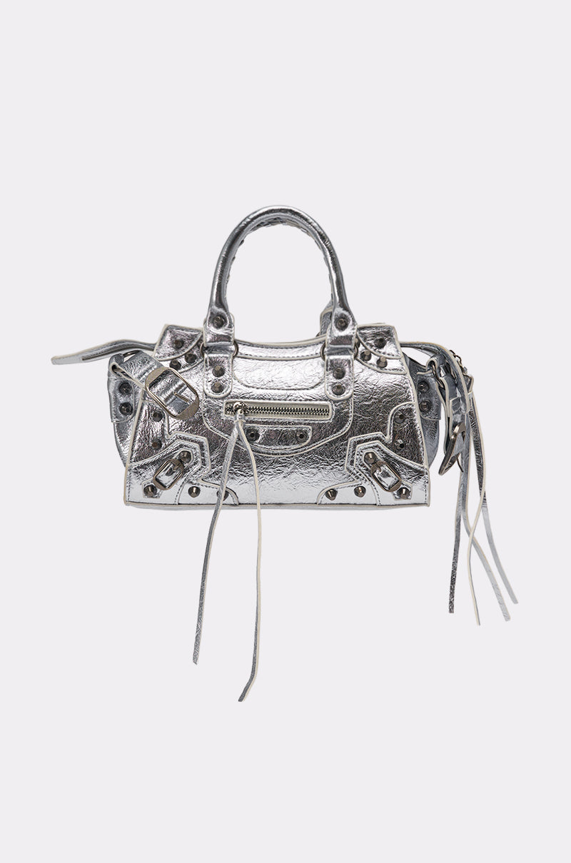 The TO THE CITY PURSE is a metallic silver faux leather mini bag with top handles, a front zip pocket, decorative buckles, long leather tassels, and a removable shoulder strap, shown against a light gray background. 3